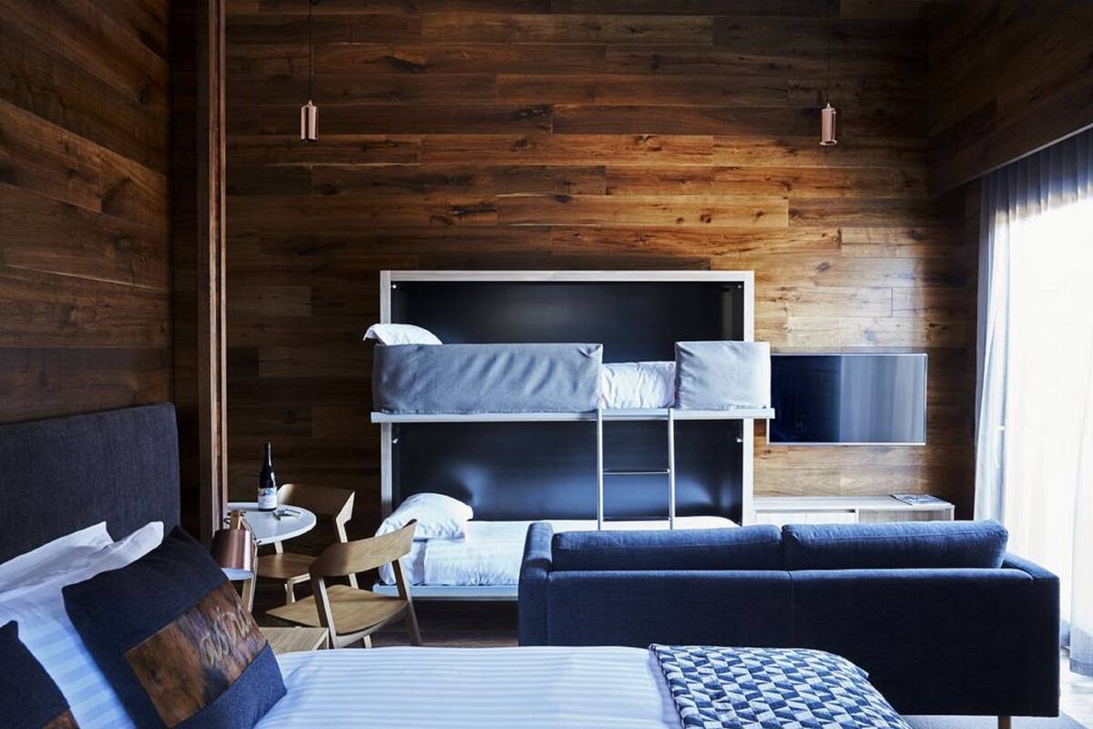 A modern hotel room with a wooden interior, a bunk bed, a sofa, a TV, and a small table with chairs.