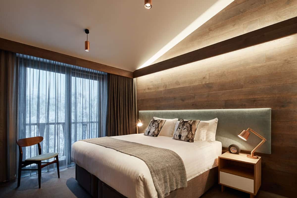 A modern hotel room with a large bed, a wooden headboard, a nightstand with a lamp, and a chair.