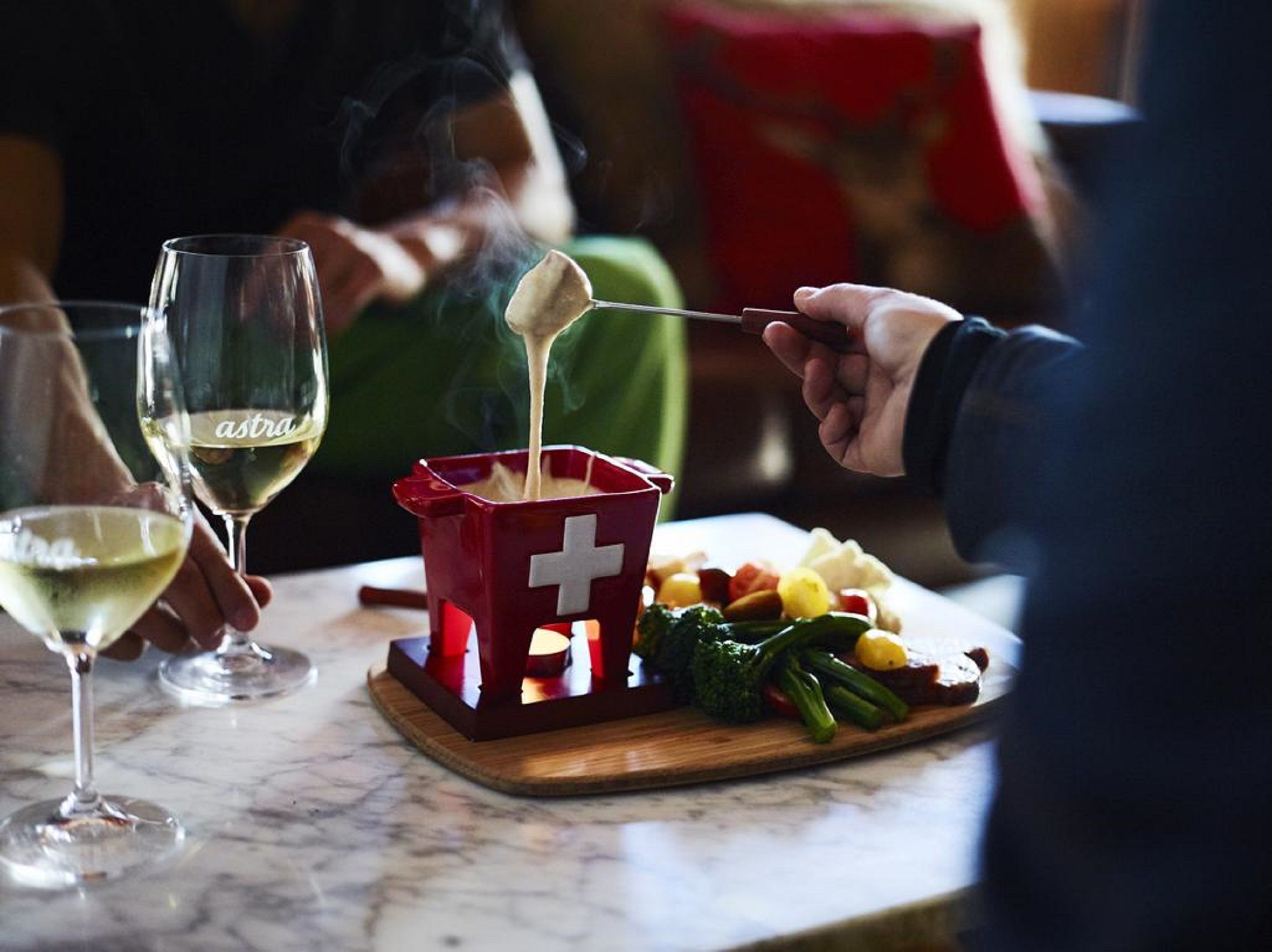 A dining table with a fondue pot, a wooden board with vegetables, and two glasses of white wine.
