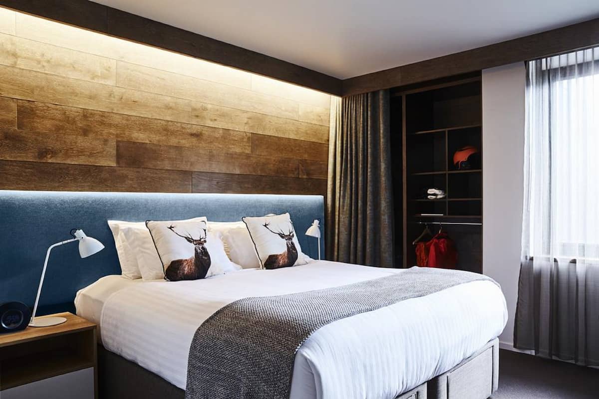 A modern hotel room with a large bed, a wooden headboard, a nightstand with a lamp, a wardrobe, and a window with curtains.