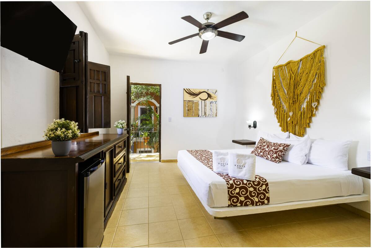 A hotel room with a bed, a ceiling fan, a wall-mounted macrame piece, and a view of the outdoors.