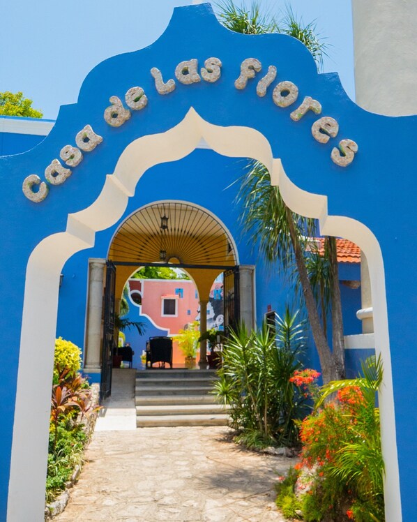 An entrance archway with the text "Casa de las flores" leading to a colorful building with a pink facade and a black chair outside. - Hotel Casa De Las Flores (Playa del Carmen)