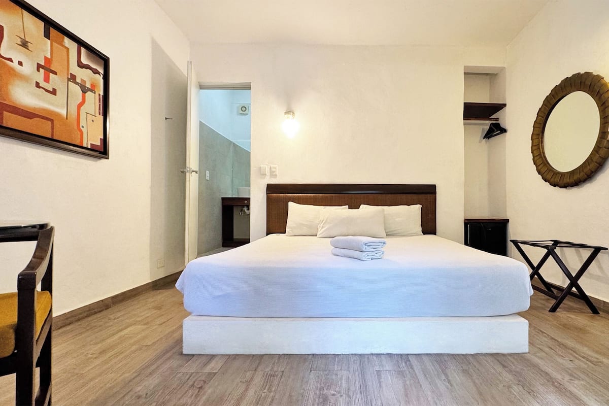 A hotel room with a large bed, a wooden floor, a desk, a chair, a mirror, and a painting on the wall.