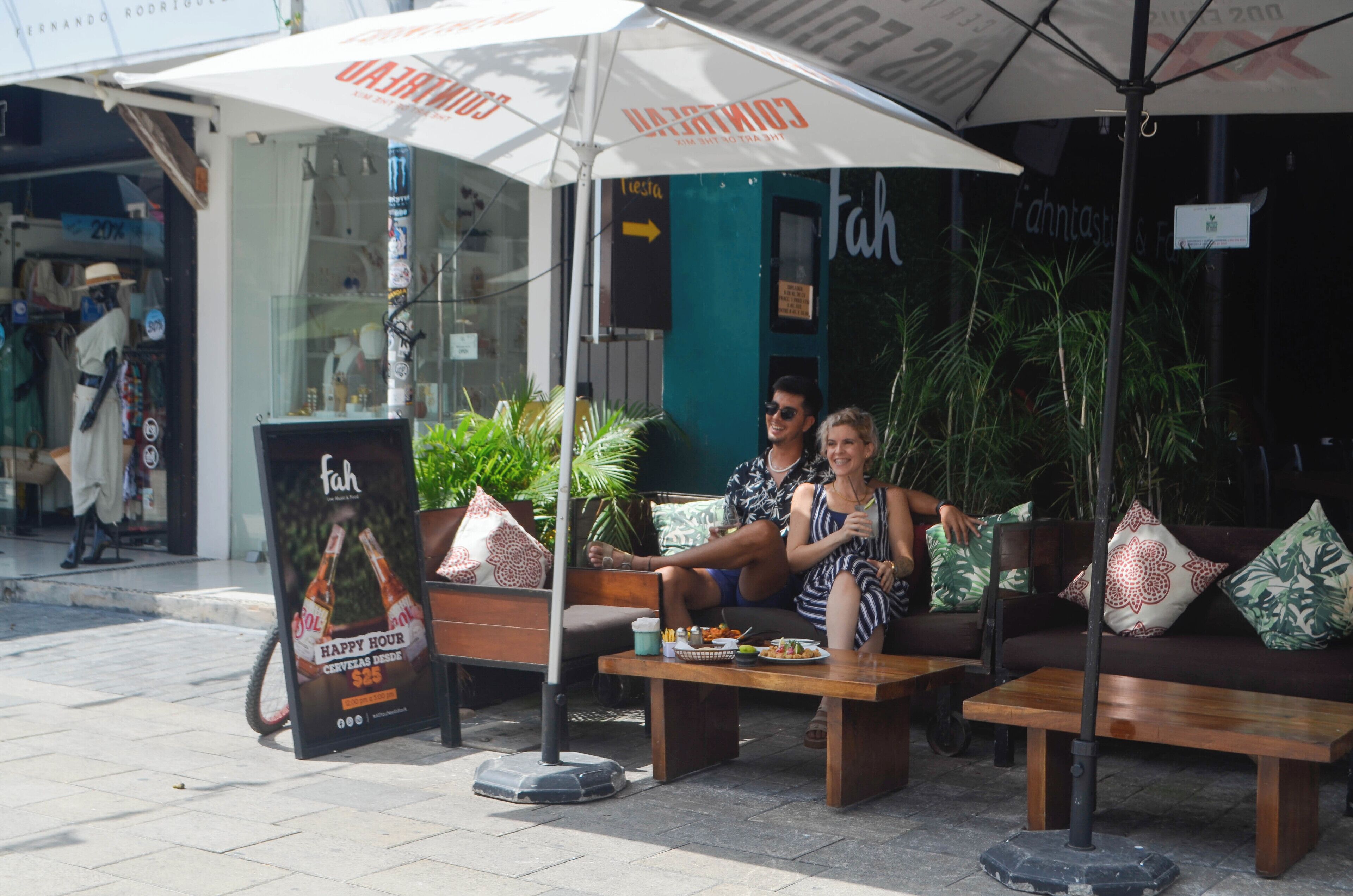 Un área de estar al aire libre con dos personas sentadas bajo sombrillas, un letrero y una tienda de fondo.