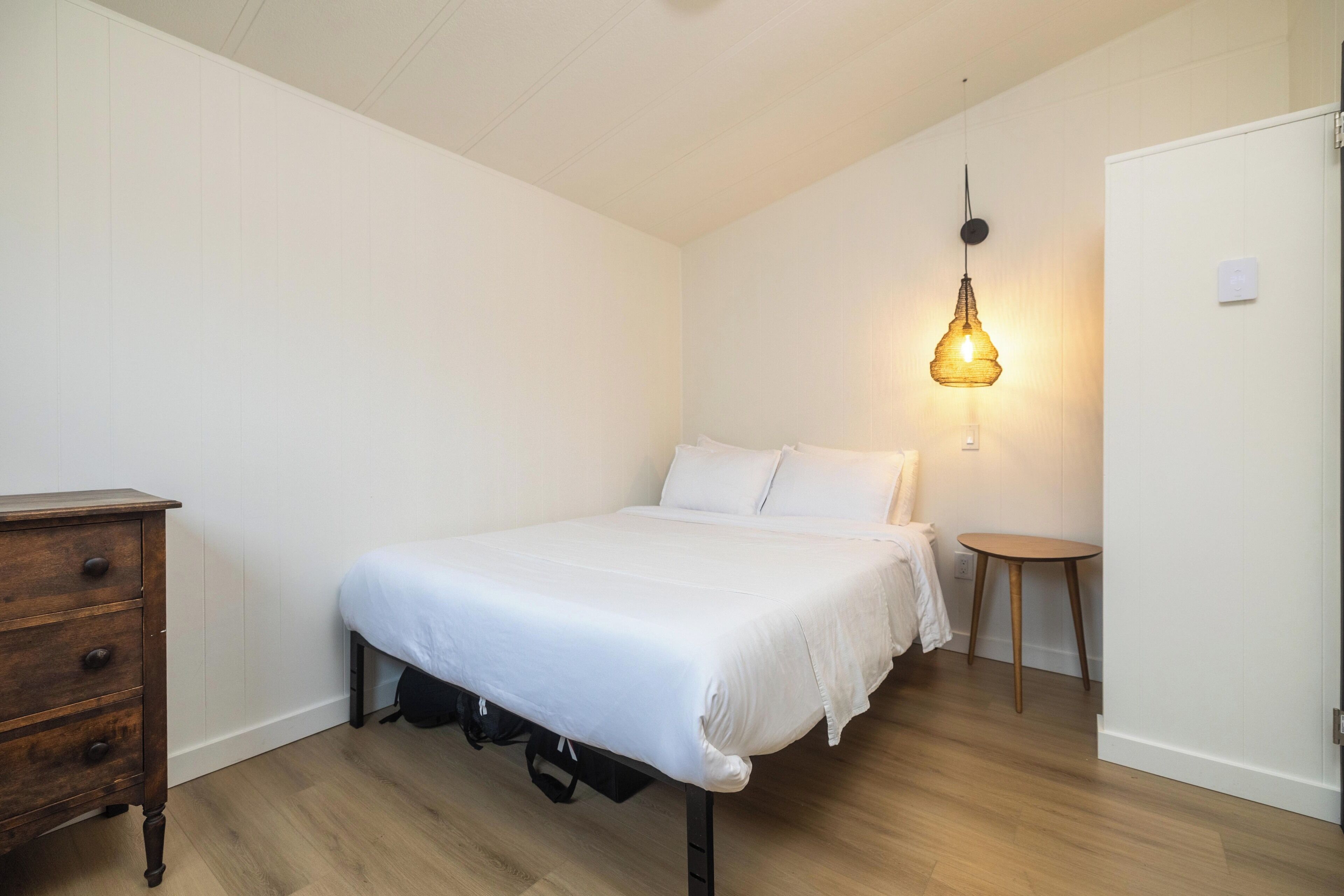 A bedroom with a bed, a dresser, a bedside table, and a hanging light fixture.