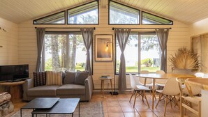 A cozy living room with a sofa, a coffee table, and a TV. There are large windows with curtains, a dining area with chairs, and a table. - Mackenzie Beach Resort (Tofino)