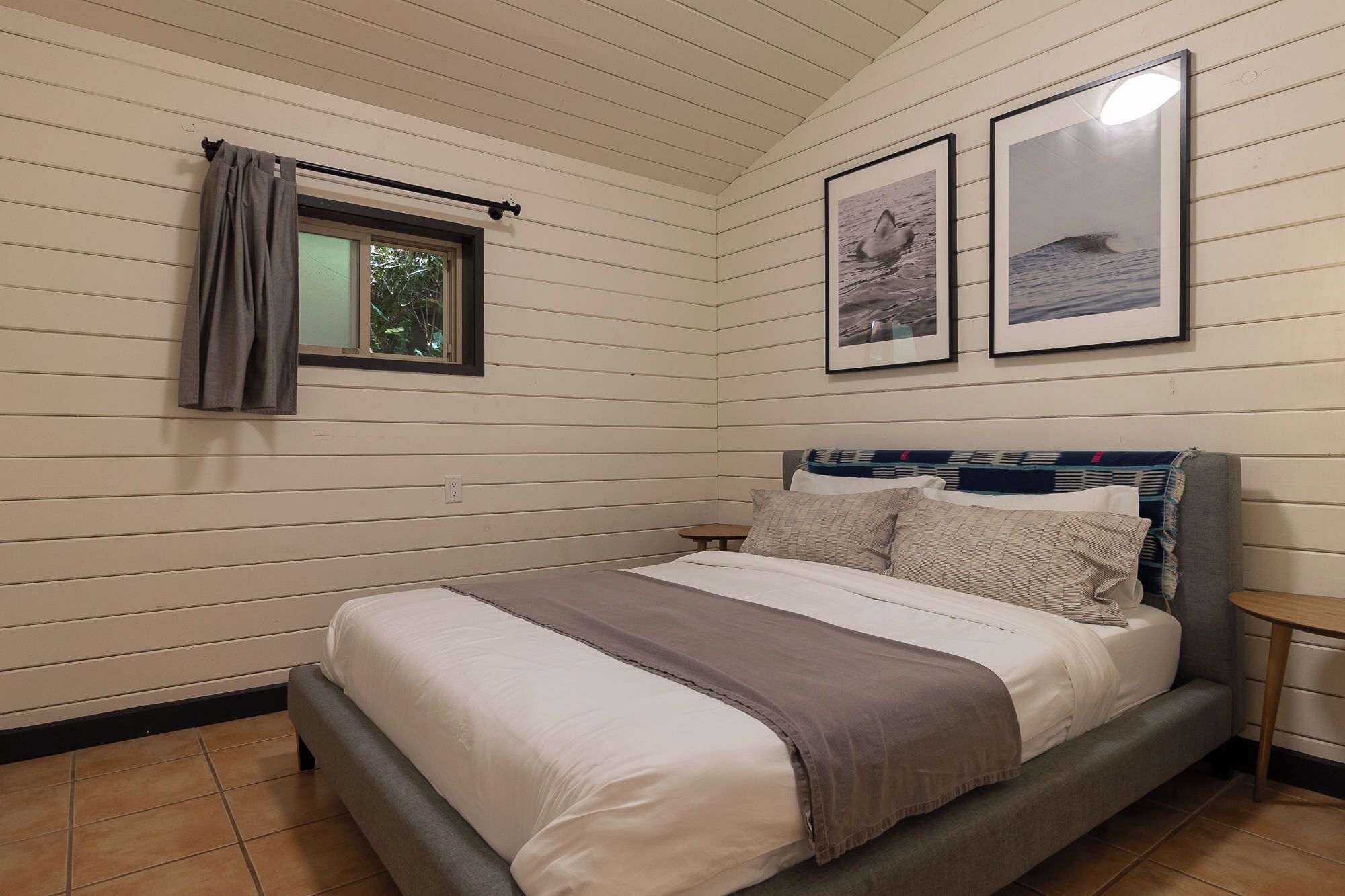 Een slaapkamer met een bed, een raam met gordijnen en twee ingelijste foto’s aan de muur.