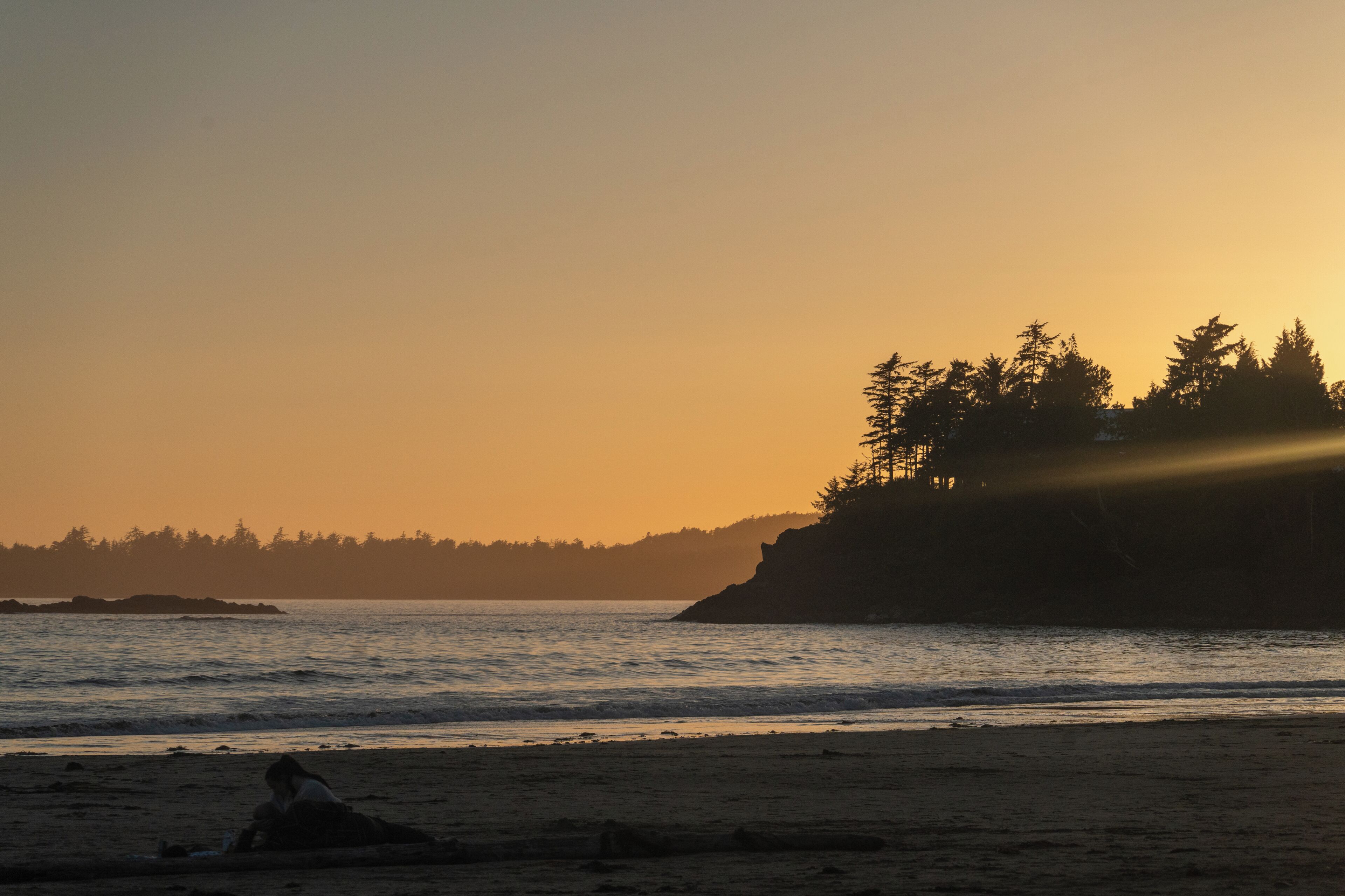 A beach with a person lying on the sand, a rocky shoreline, and trees in the background.