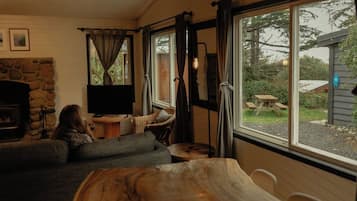A cozy living room with a stone fireplace, a television, and a large window overlooking a garden.