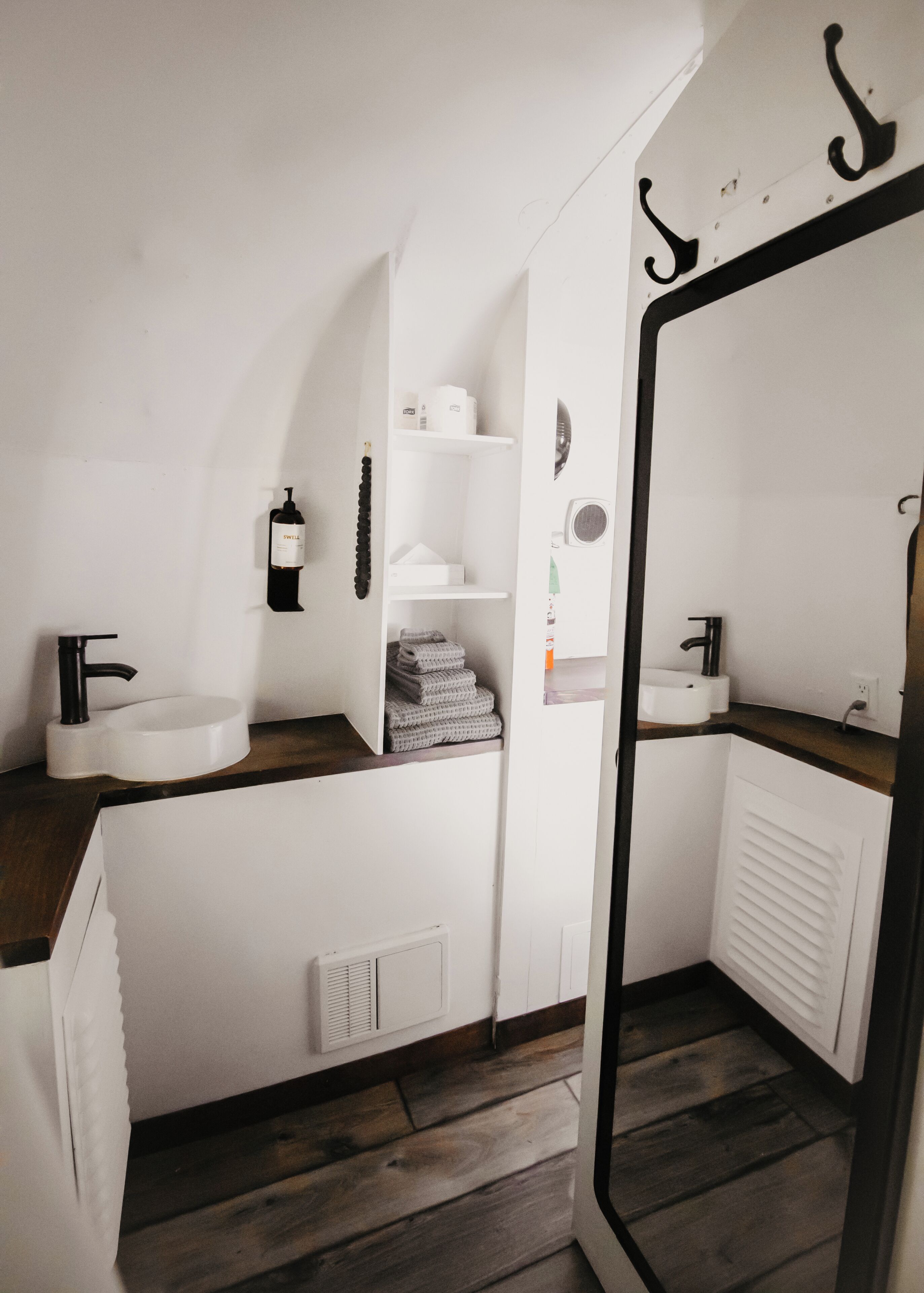 A modern bathroom with a white sink, black faucet, and a large mirror. There are shelves with toiletries and a towel rack.