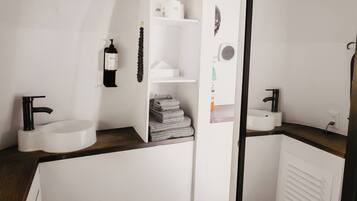 A modern bathroom with a white sink, black faucet, and a large mirror. There are shelves with toiletries and a towel rack.