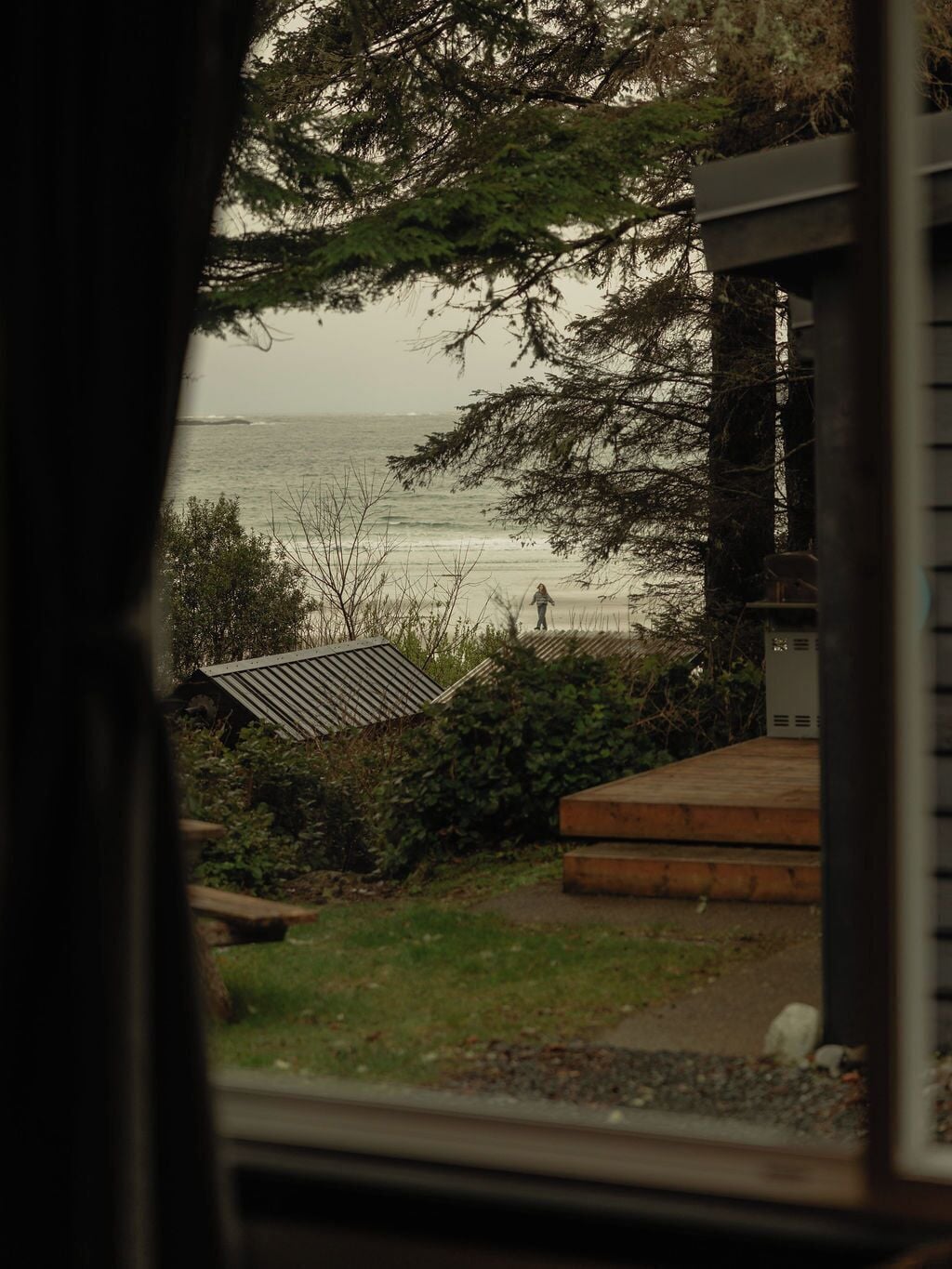 A view through a window of a coastal property with a wooden deck, a metal roof structure, and a person walking along the beach.