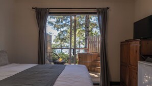 A bedroom with a bed, a TV, a wooden cabinet, and a balcony with a view of trees and a body of water. - Mackenzie Beach Resort (Tofino)
