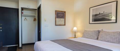 A hotel room with a bed, a desk, a chair, a coat rack, a framed picture, and a wall-mounted light.