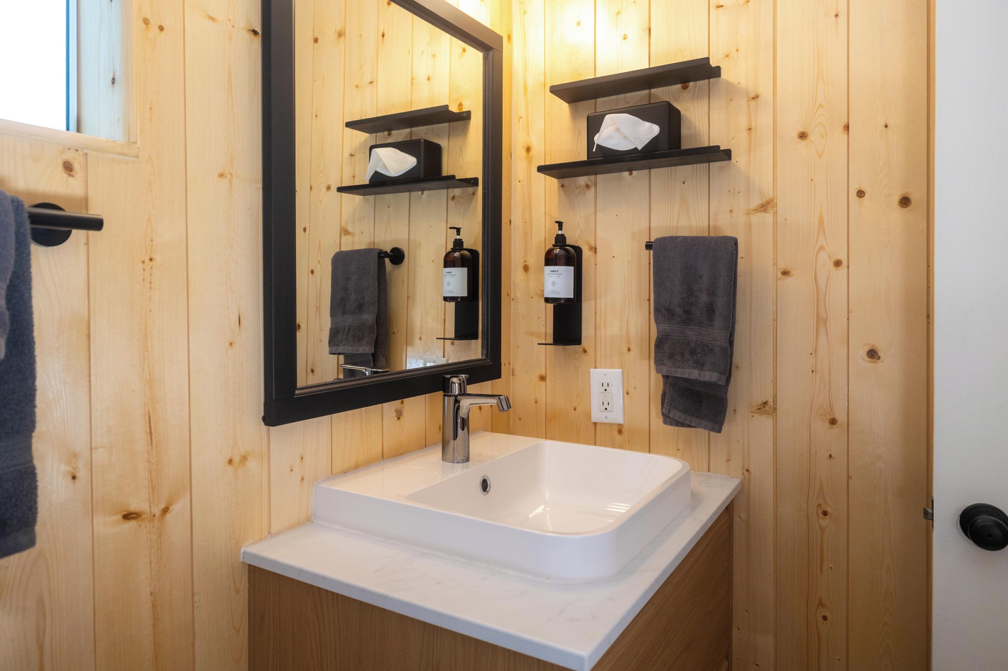 A bathroom with a white sink, a mirror, and wooden walls. There are two towels hanging on hooks and a soap dispenser on the wall.