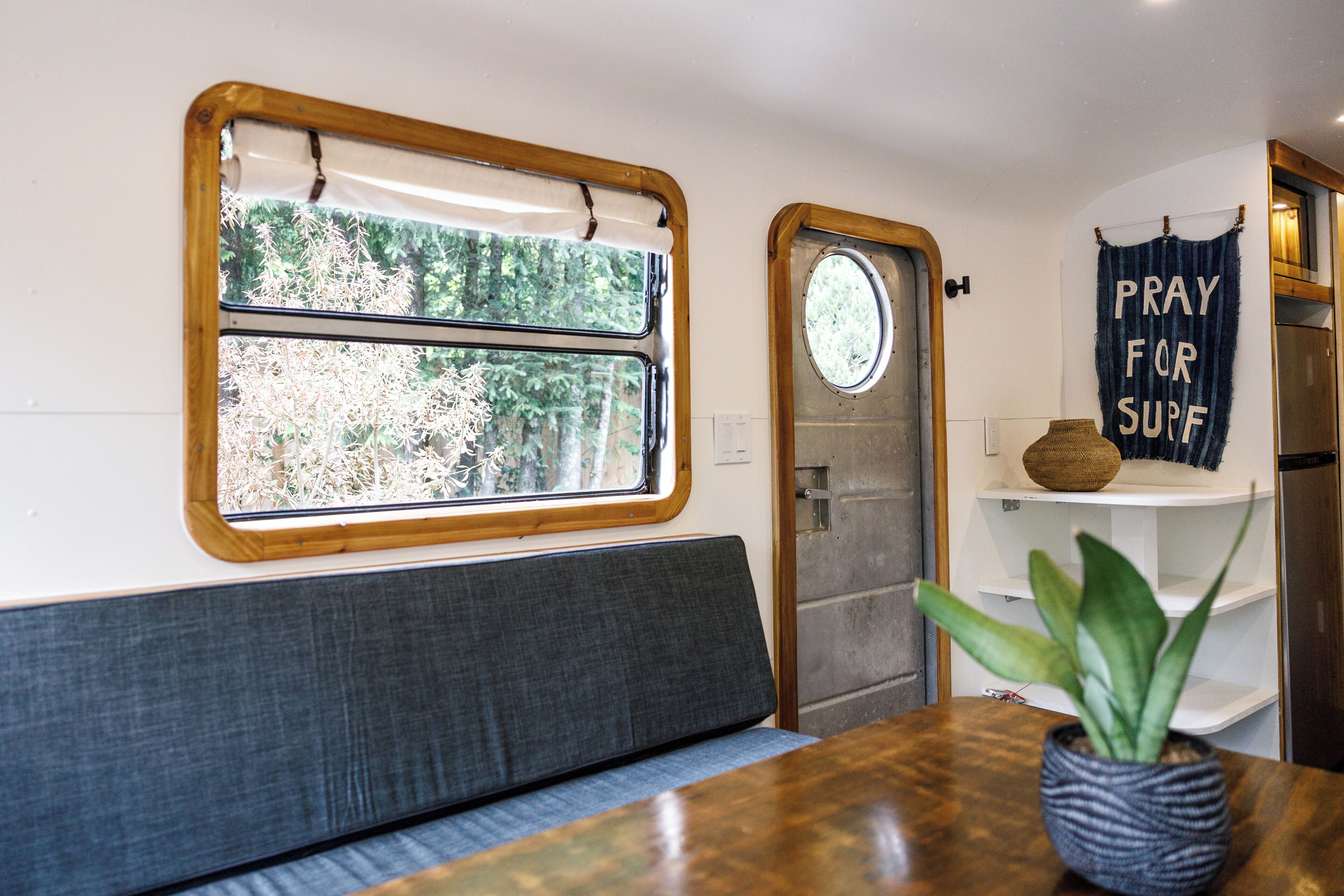 A compact RV interior with a window, a door, a bench, a shelf with a sign, and a potted plant.