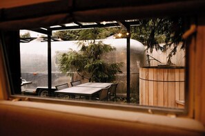 Uitzicht door een raam op een eethoek met een tafel, stoelen, een houten hek en een grote watertank op de achtergrond.