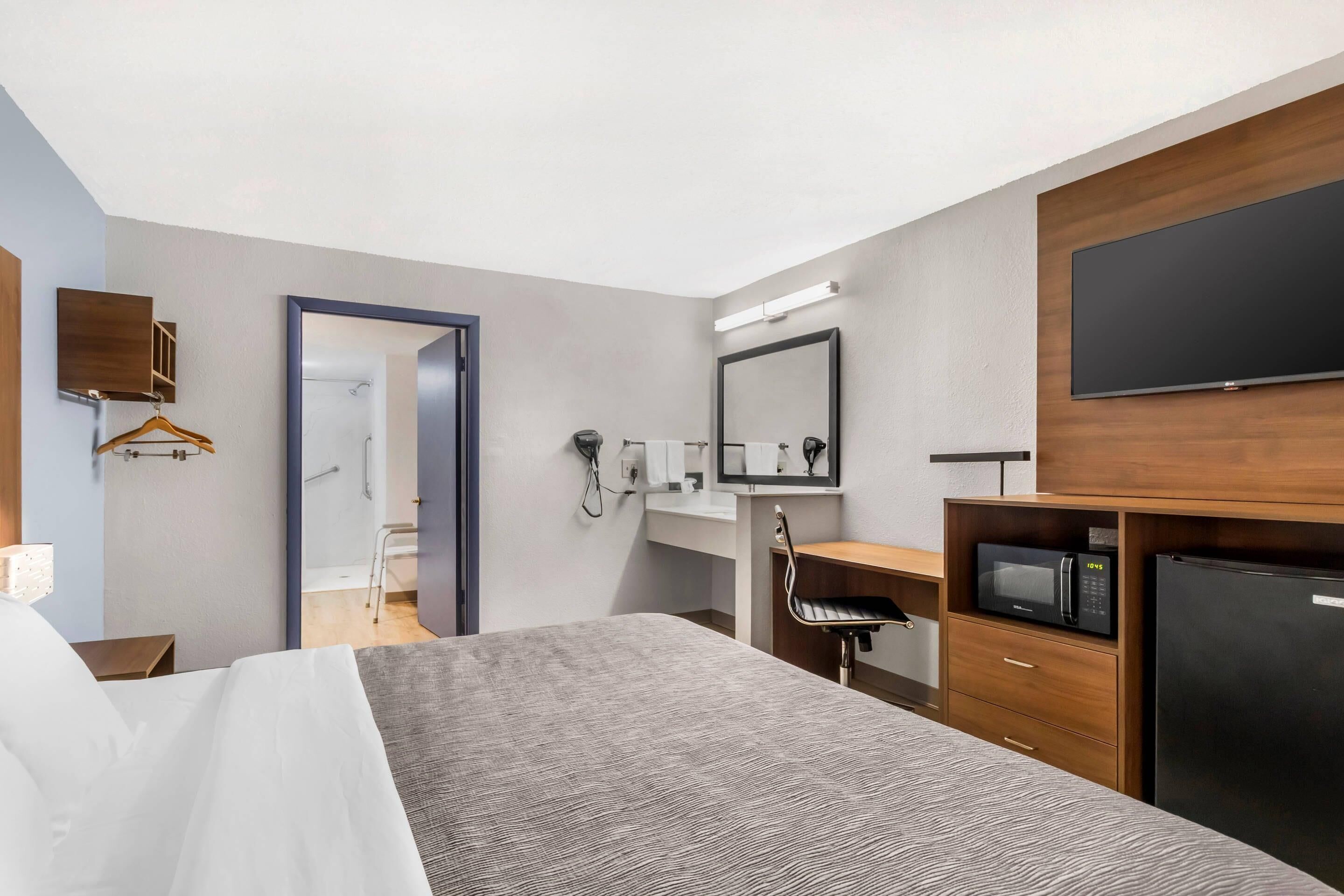 A hotel room with a bed, a desk, a microwave, a refrigerator, a TV, and a bathroom visible through an open door.