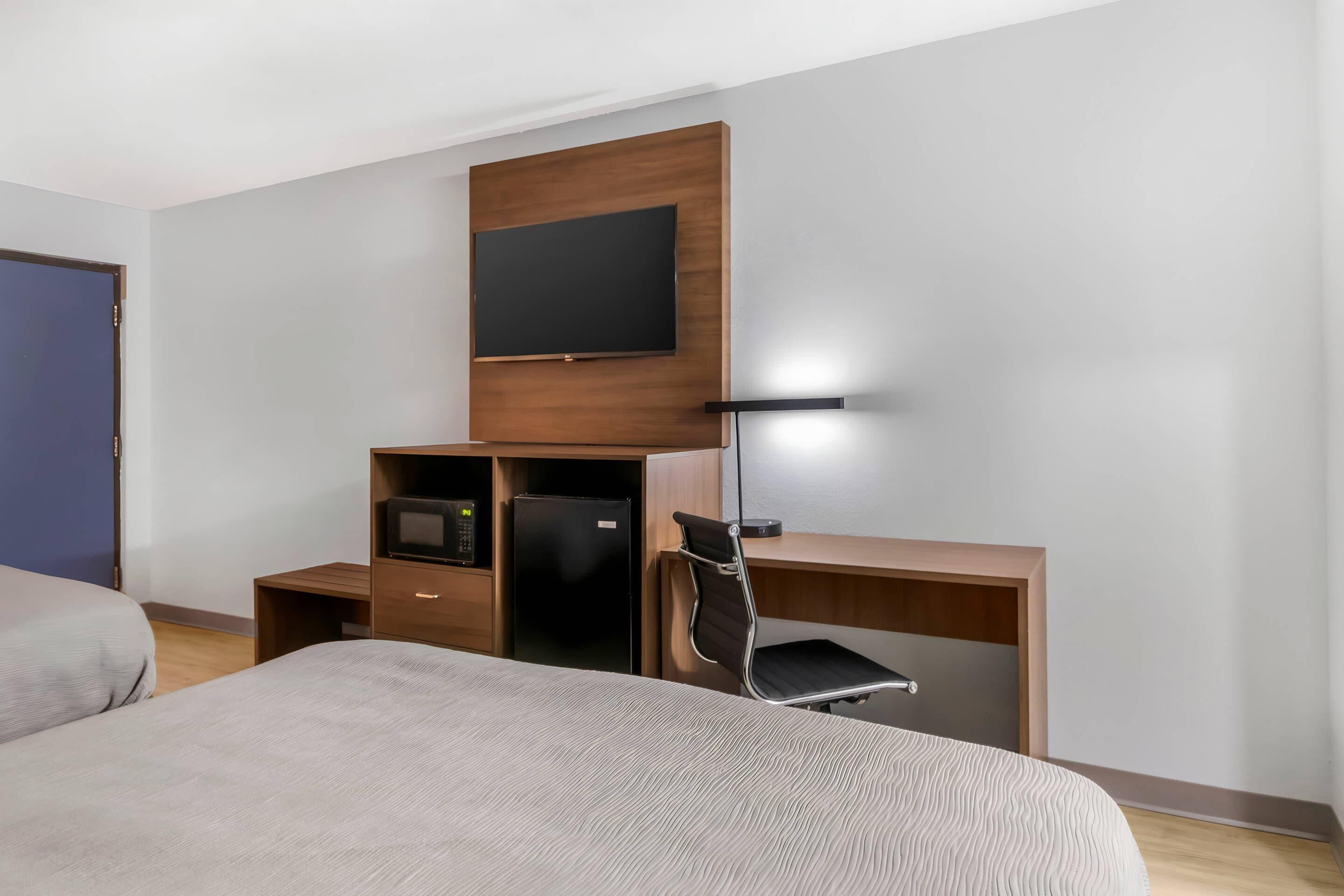 A modern hotel room with a large bed, a desk with a chair, a flat-screen TV mounted on a wooden panel, a refrigerator, and a microwave.