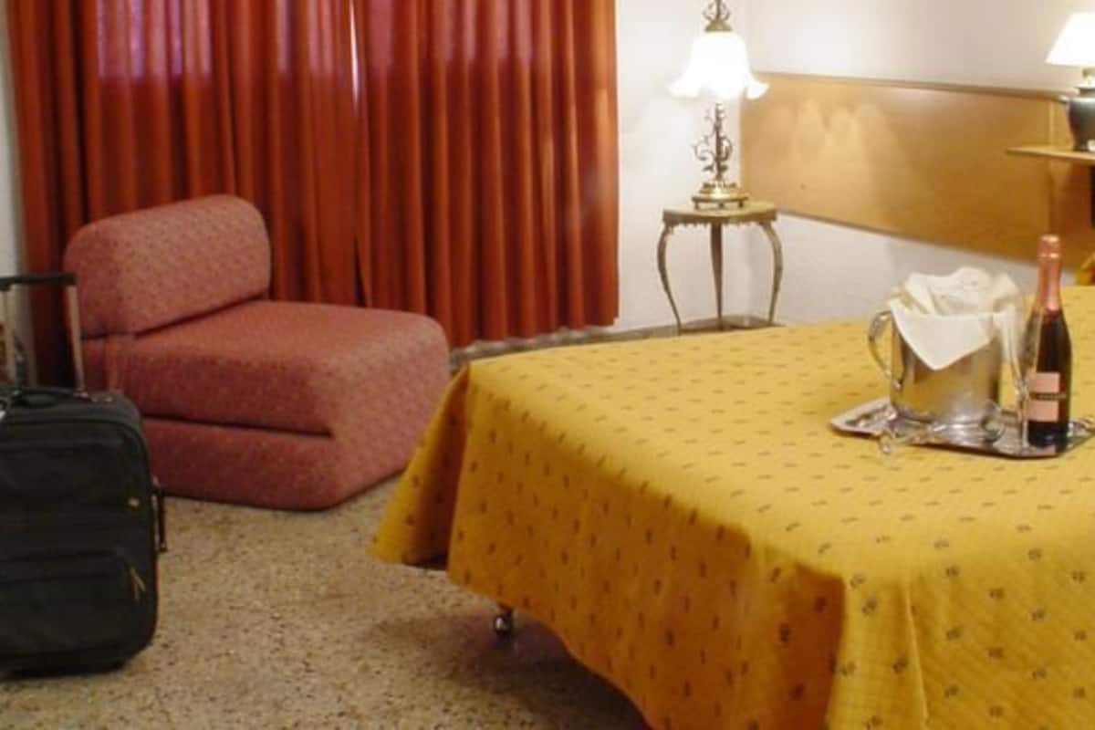 A hotel room with a bed, a chair, a suitcase, and a bottle of wine on the bed.