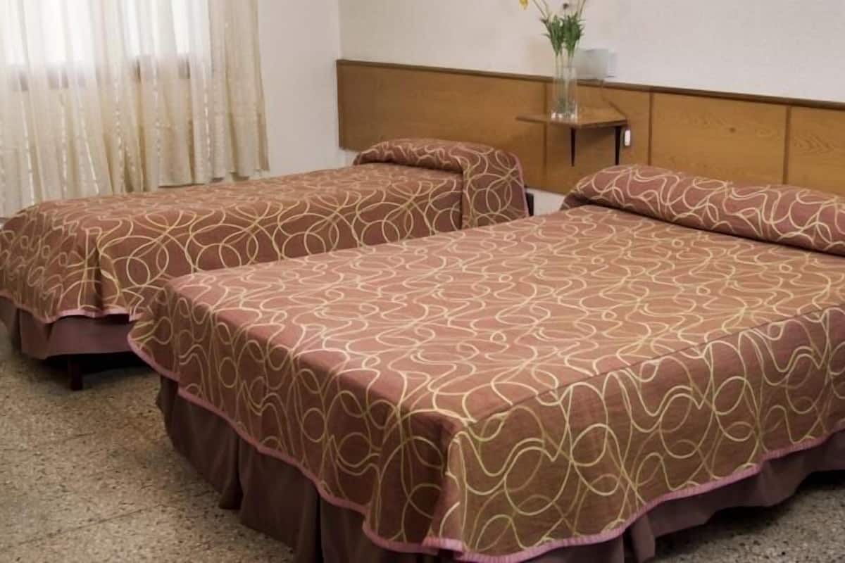 A hotel room with two beds, a wooden headboard, a small bedside table with a vase, and a window with curtains.