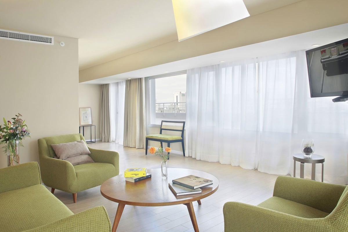 A modern living room with green upholstered chairs, a round wooden coffee table, and a view of the city through large windows.
