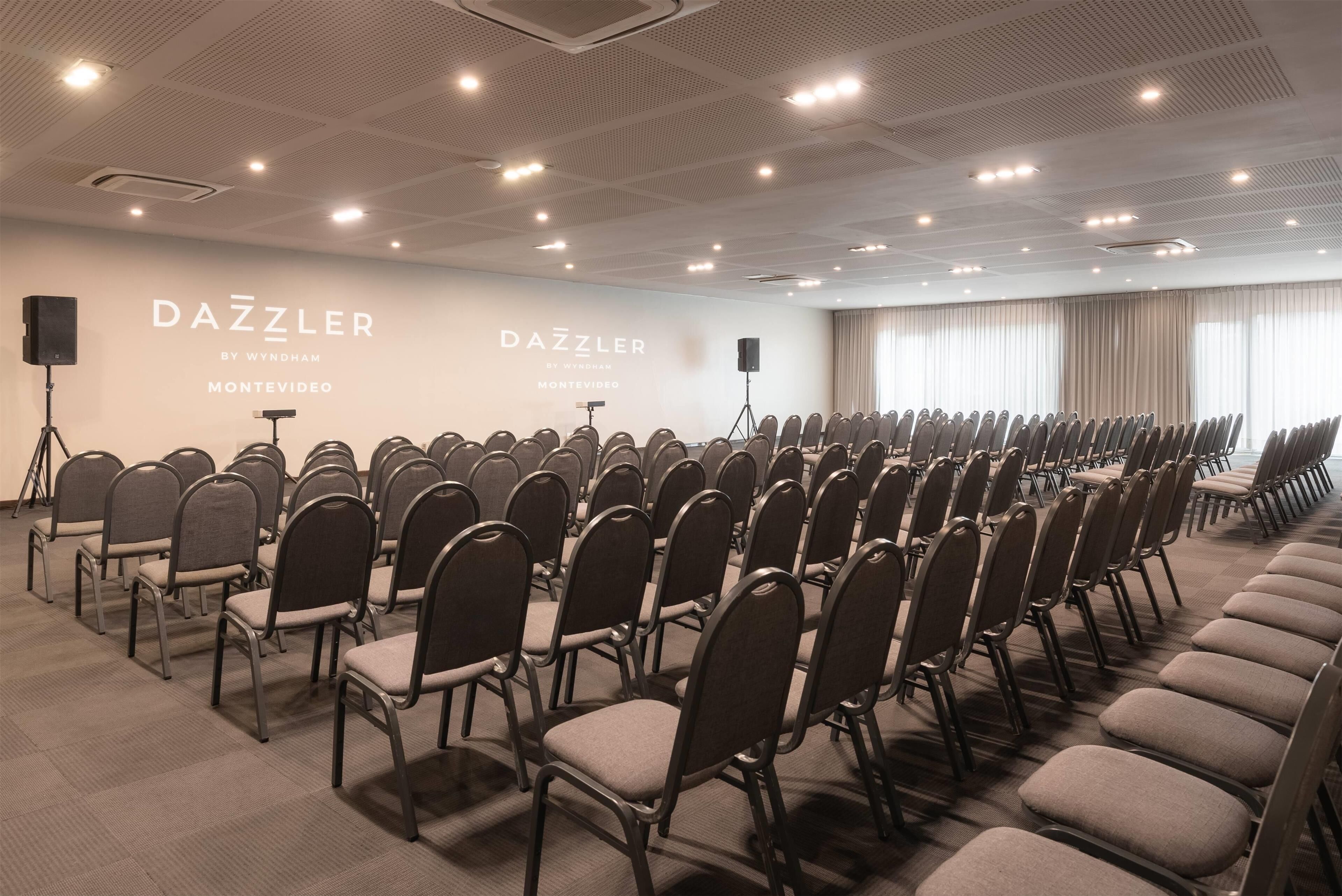 Une salle de conférence avec des rangées de chaises, une scène où se trouve un orateur, et une grande fenêtre avec des rideaux.
