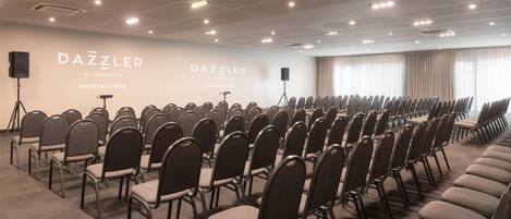 Une salle de conférence avec des rangées de chaises, une scène où se trouve un orateur, et une grande fenêtre avec des rideaux.