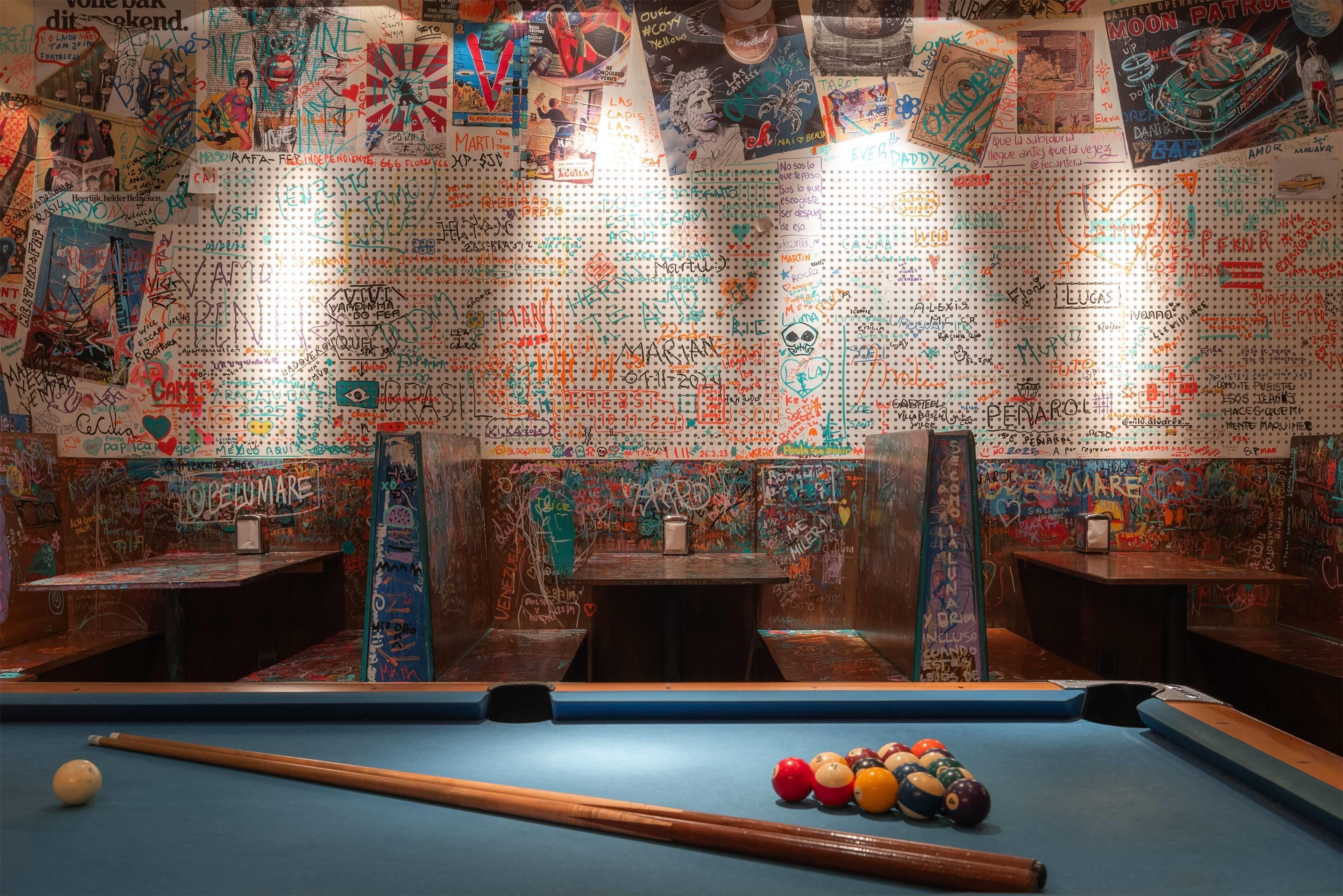 A pool table with a blue felt surface, surrounded by walls covered in colorful graffiti and writings.