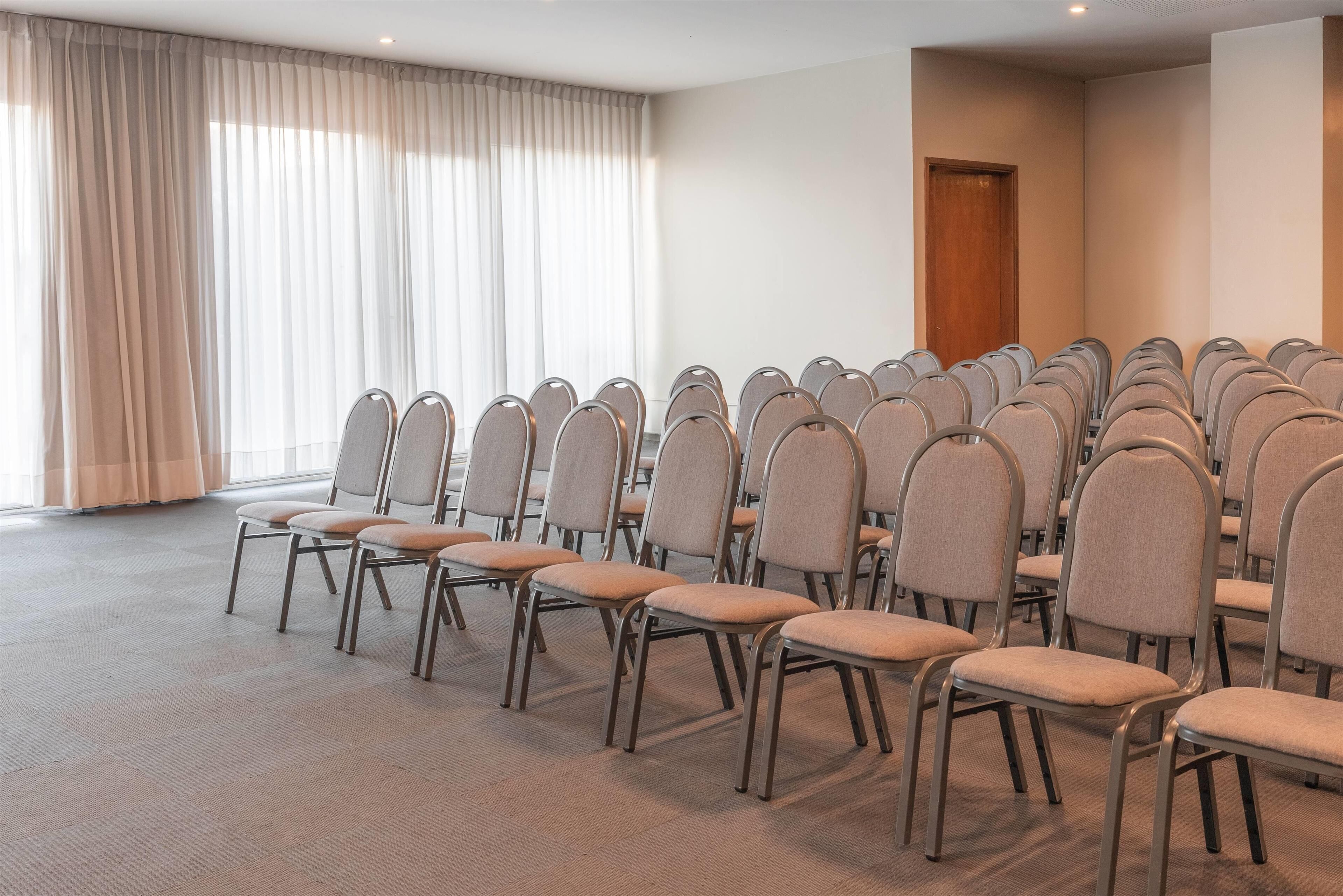 A conference room with rows of chairs, a large window with curtains, and a door.