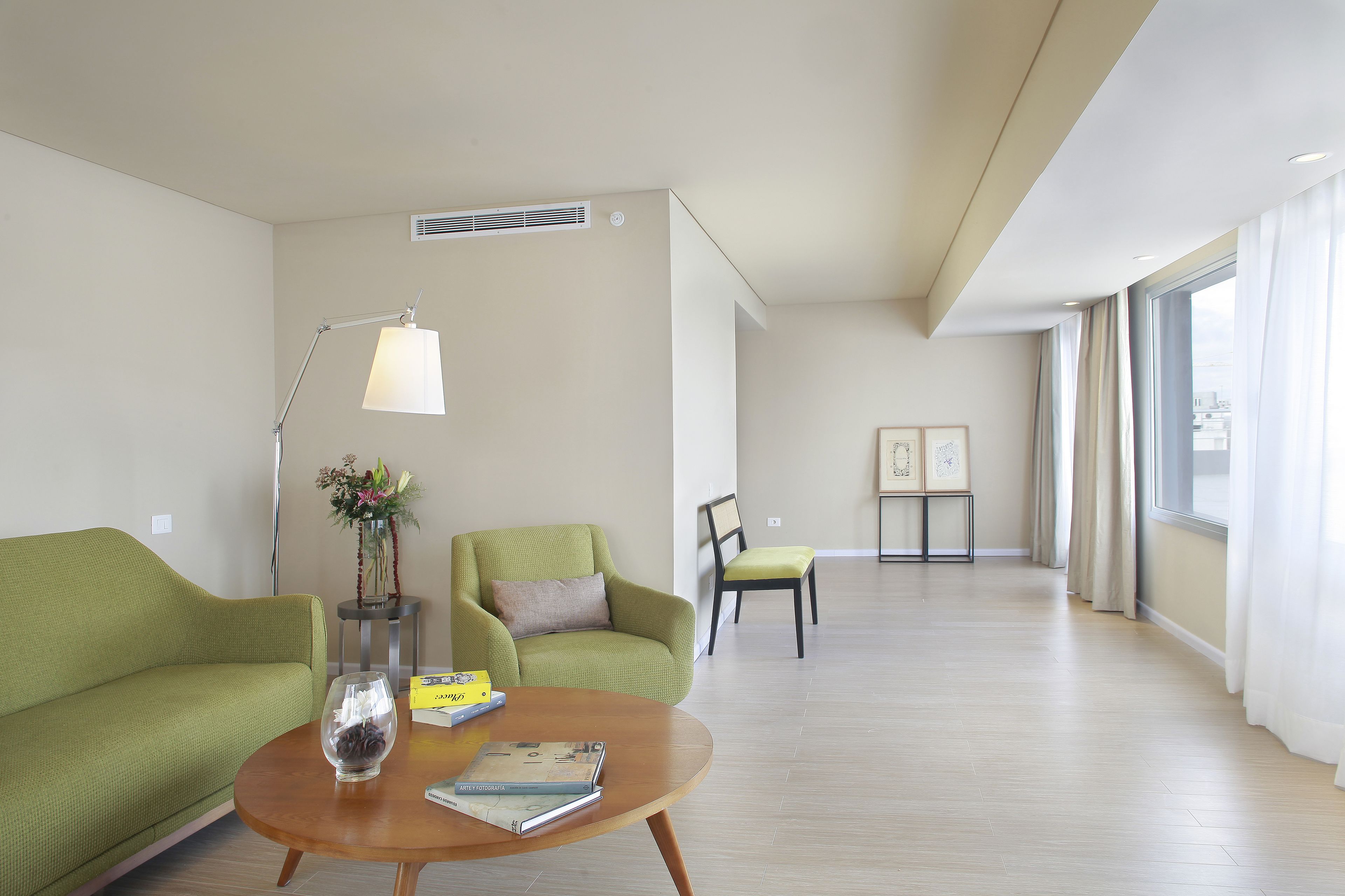 Una sala de estar moderna con un sofá verde, una mesa de centro de madera y una lámpara de pie. Hay una mesita con un florero y un libro. La habitación tiene ventanas amplias con cortinas y vistas a la ciudad.