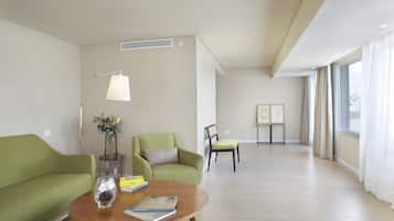 Una sala de estar moderna con un sofá verde, una mesa de centro de madera y una lámpara de pie. Hay una mesita con un florero y un libro. La habitación tiene ventanas amplias con cortinas y vistas a la ciudad.