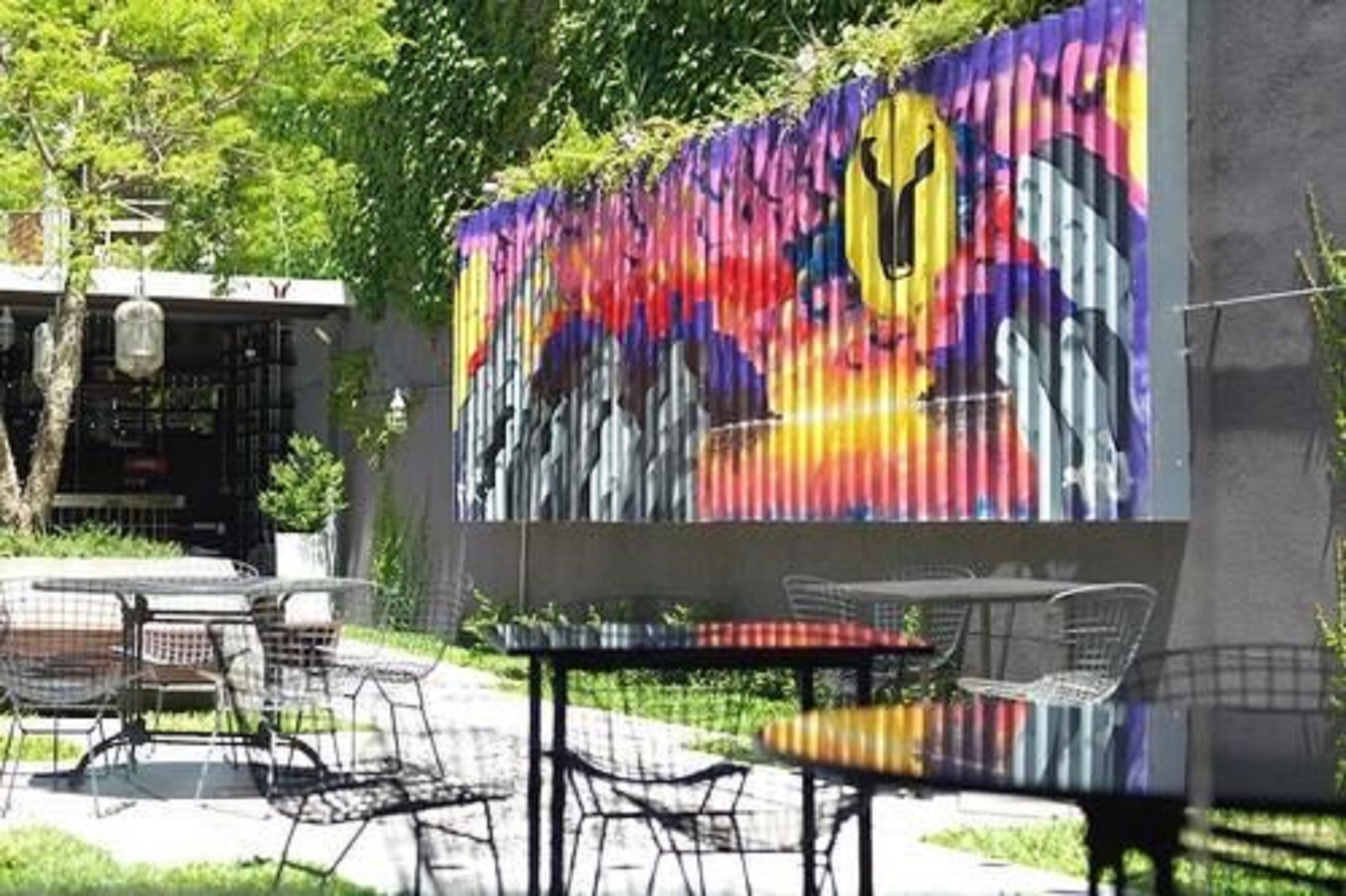 Un espace extérieur aménagé avec des chaises et une table en métal, une fresque colorée sur le mur et un bâtiment dont l’entrée est bien visible.