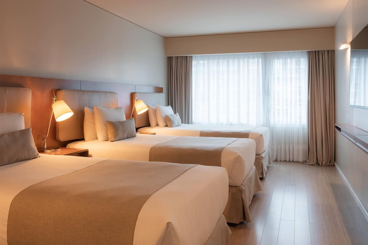 A hotel room with two beds, each with a bedside table and lamp, a large window with curtains, and a wooden floor.
