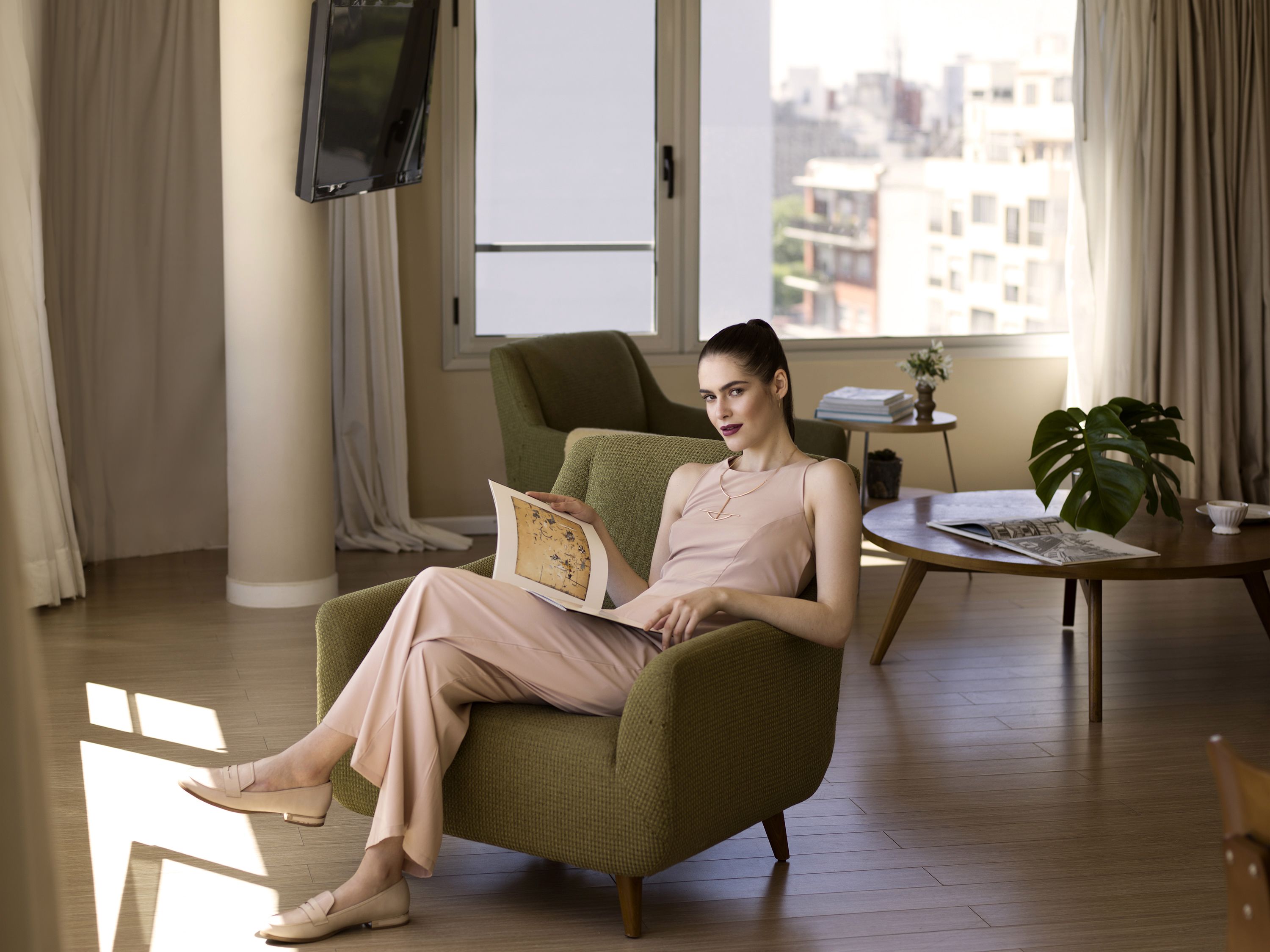 Eine Person sitzt in einem grünen Sessel in einem Zimmer mit Blick auf die Stadt, an der Wand ist ein Fernseher angebracht, und ein kleiner Tisch mit Pflanze und Büchern steht daneben.