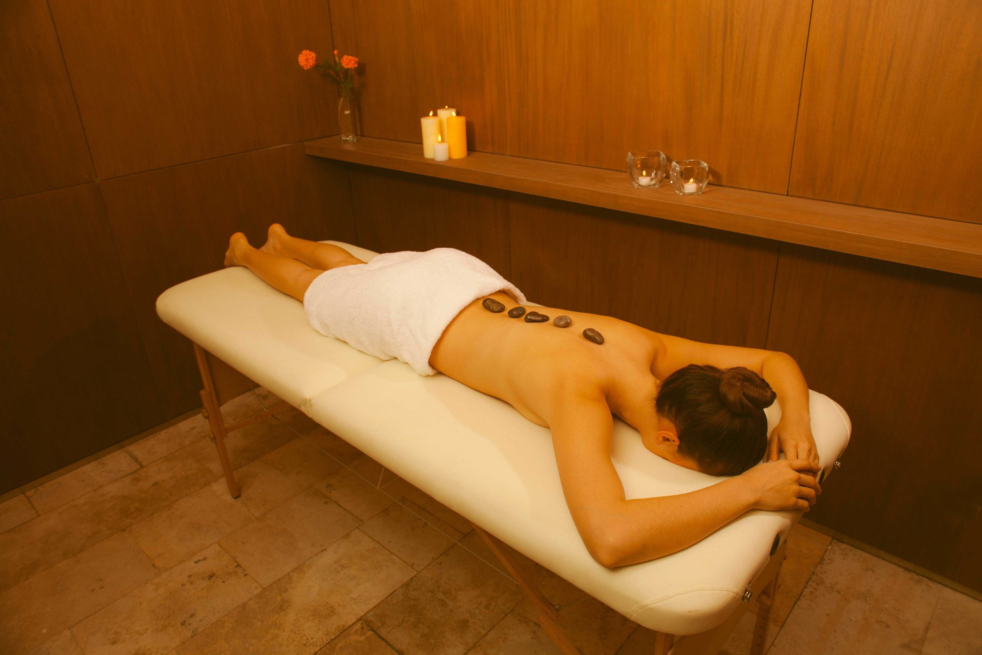 Une personne reçoit un massage du dos sur une table de massage blanche, dans une pièce aux murs lambrés, où l’on aperçoit une étagère avec des bougies et un vase de fleurs.
