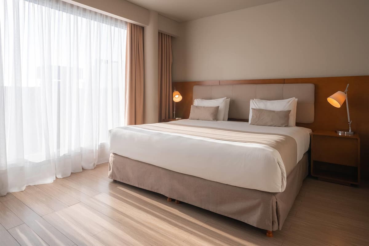 A hotel room with a large bed, two bedside lamps, a wooden headboard, and sheer curtains.