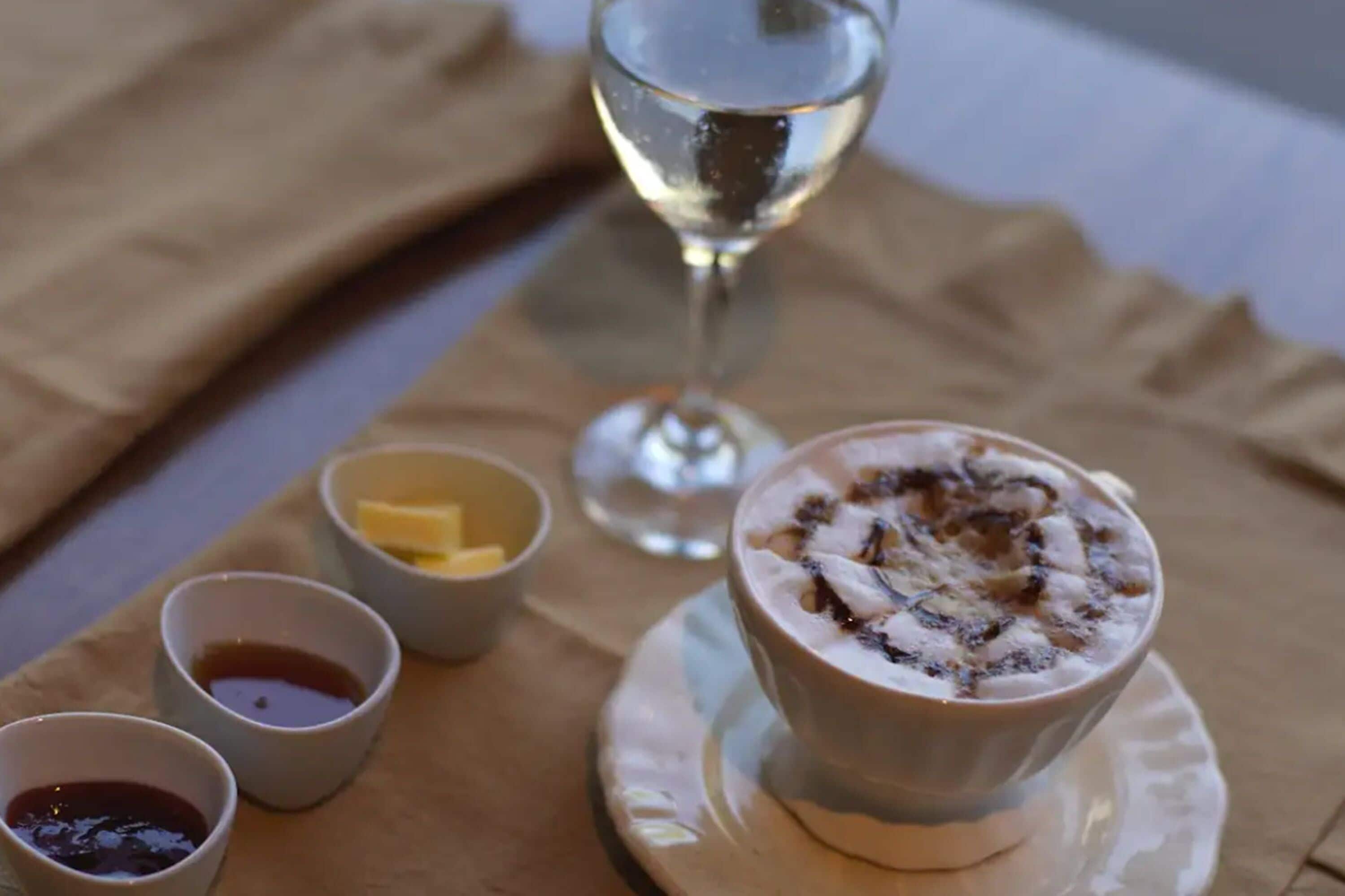A dessert with whipped cream and chocolate shavings, accompanied by small bowls of butter and sauces, and a glass of white wine on a table.