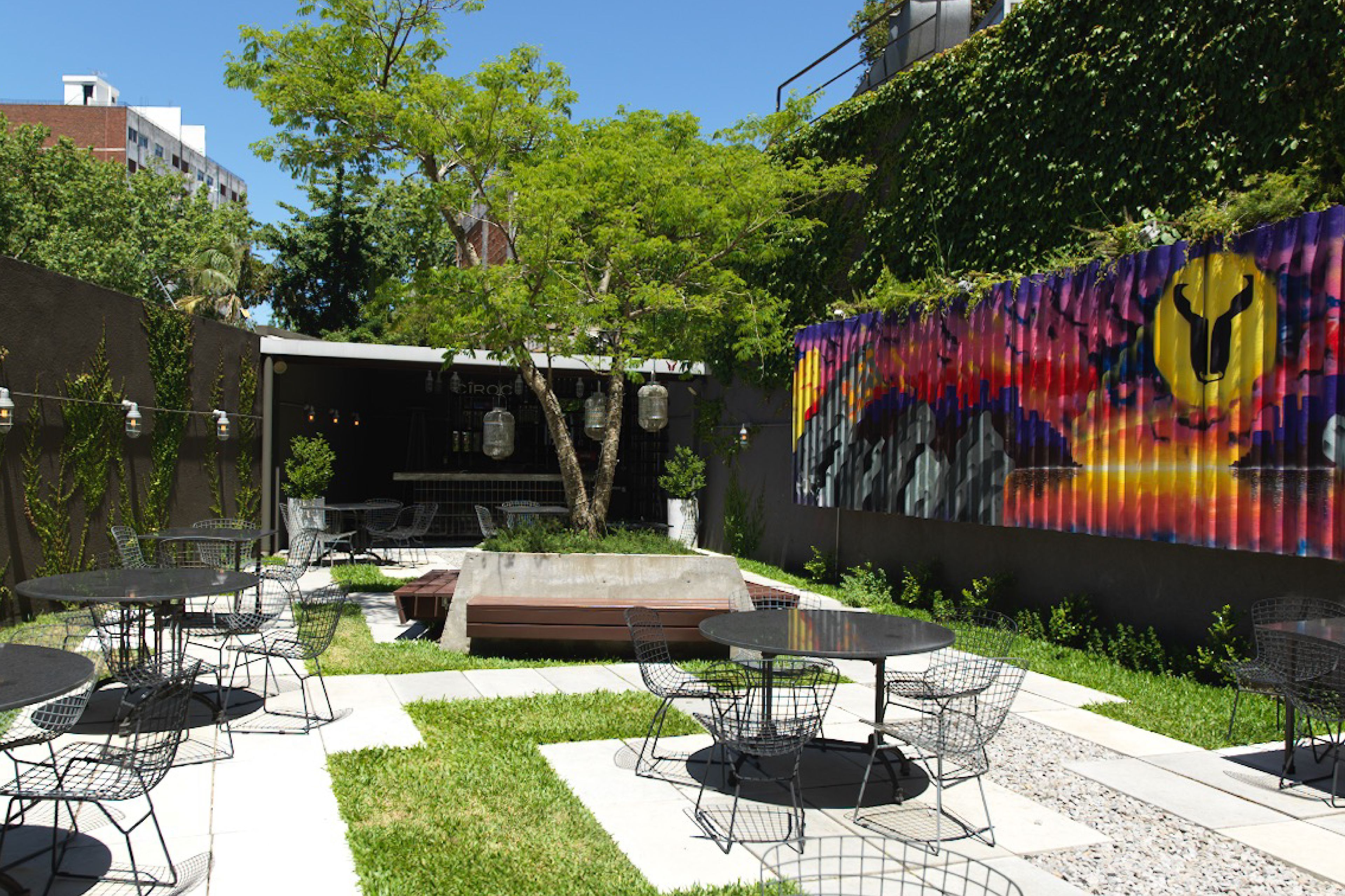 Un patio al aire libre con mesas y sillas redondas, un banco de hormigón y un mural en la pared.