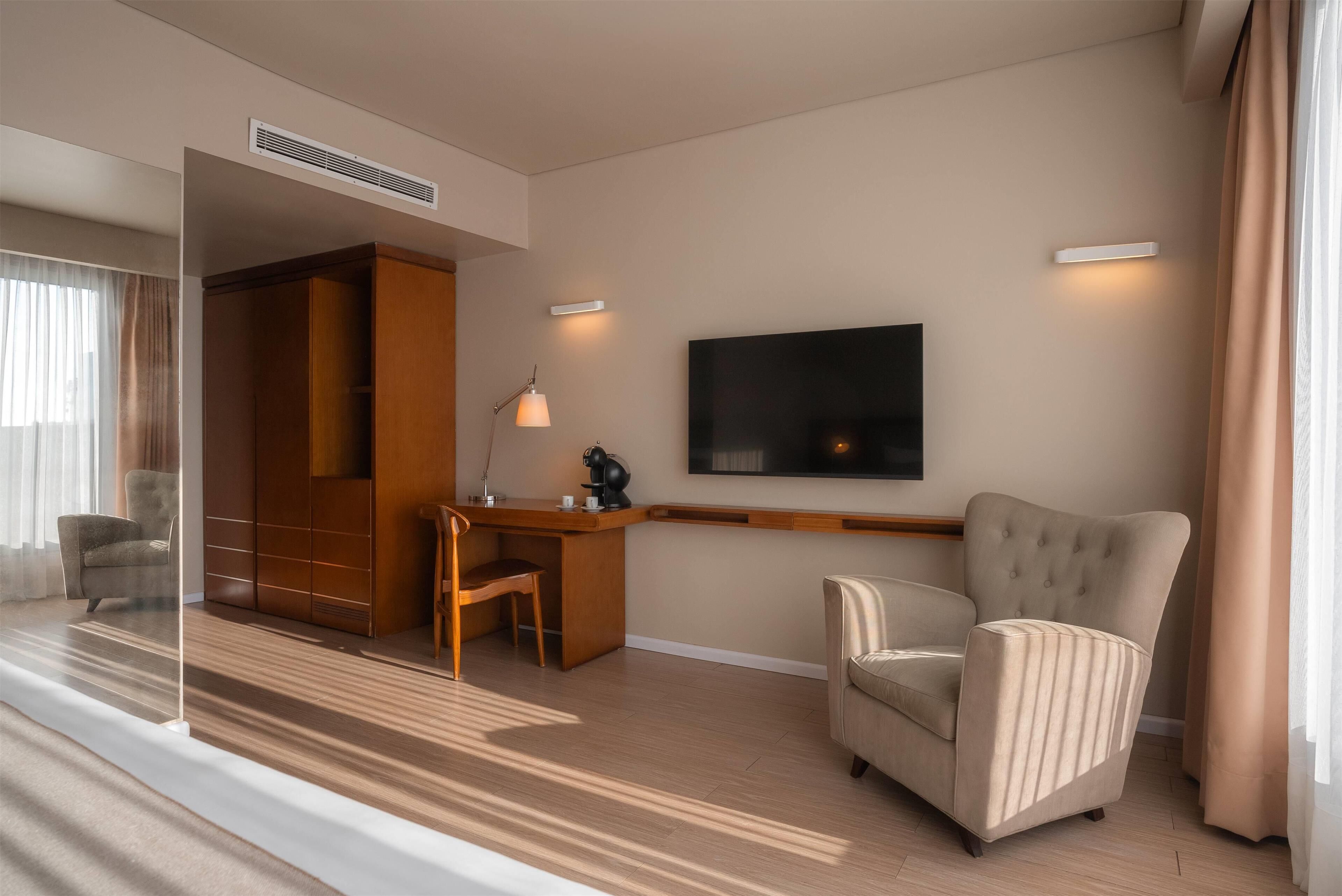 Una habitación de hotel moderna con un escritorio de madera, un sillón cómodo, un televisor de pantalla plana en la pared y un amplio armario.