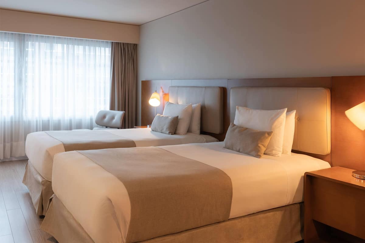 A hotel room with two beds, a large window with curtains, and a bedside lamp.