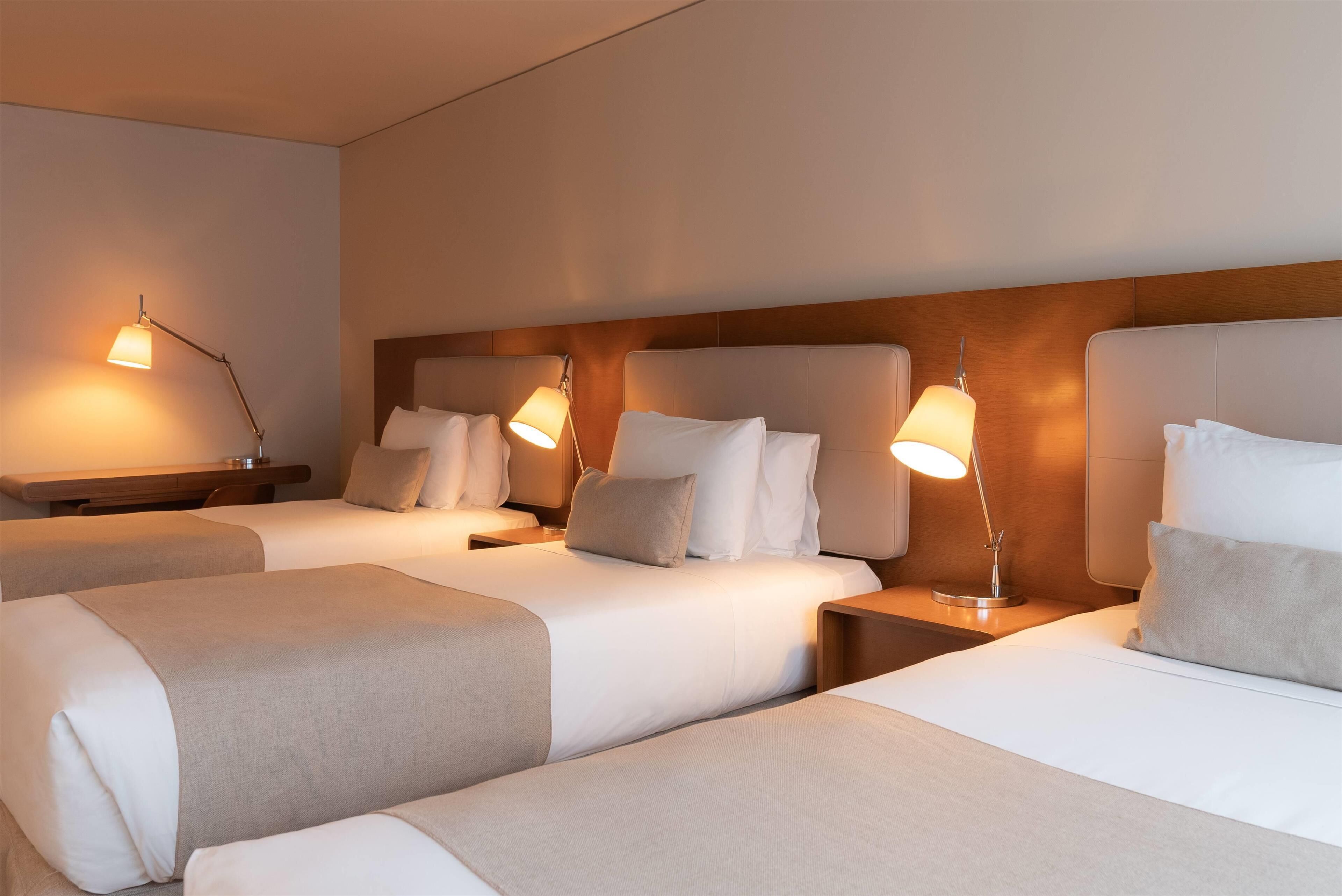 A hotel room with three beds, each with a bedside table and lamp.