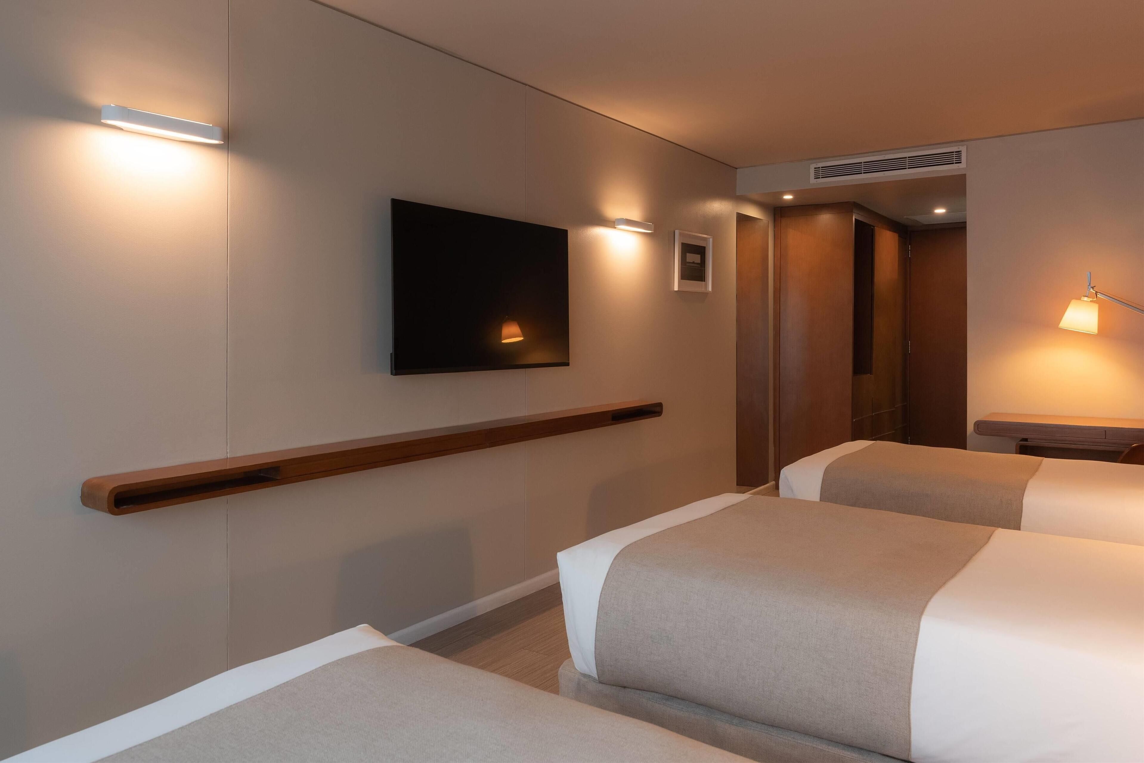 Habitación de hotel con dos camas, un televisor montado, luces empotradas en la pared y un estante de madera.