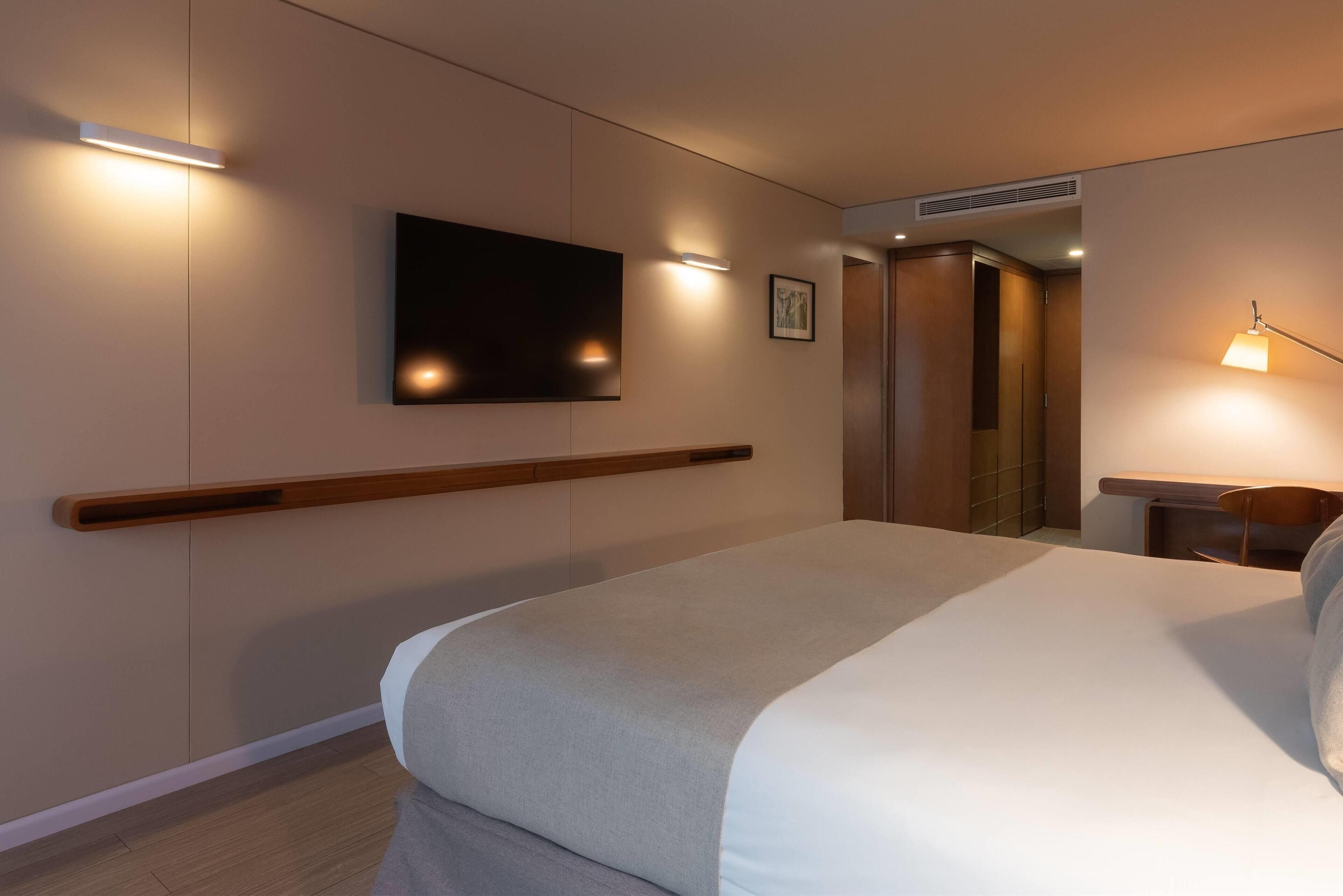 Una habitación de hotel con una cama, televisor montado, lámparas de noche y cabecero de madera.