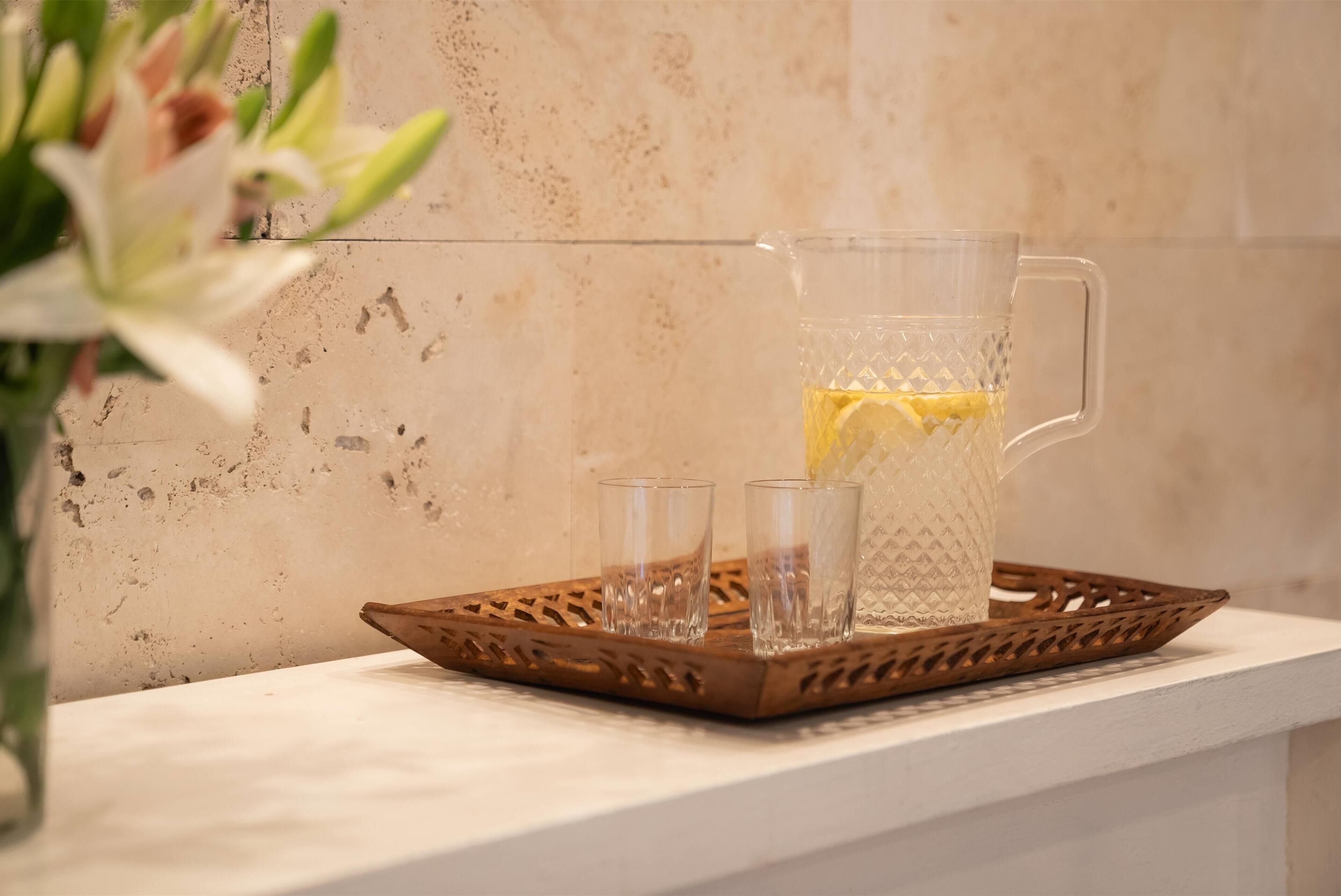 Un jarro de vidrio transparente con una rodaja de limón sobre una bandeja, junto a dos vasos pequeños, frente a una pared con textura.