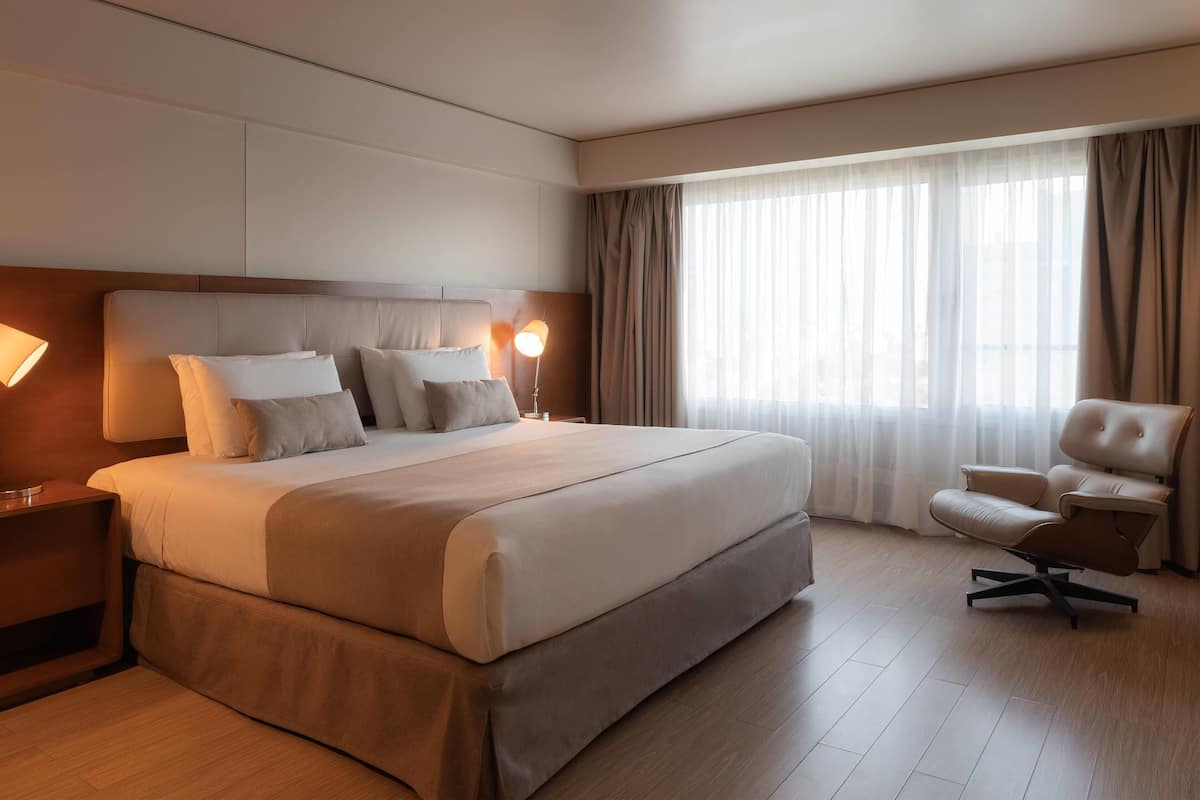 A modern hotel room with a large bed, a desk, a chair, and a window with curtains.