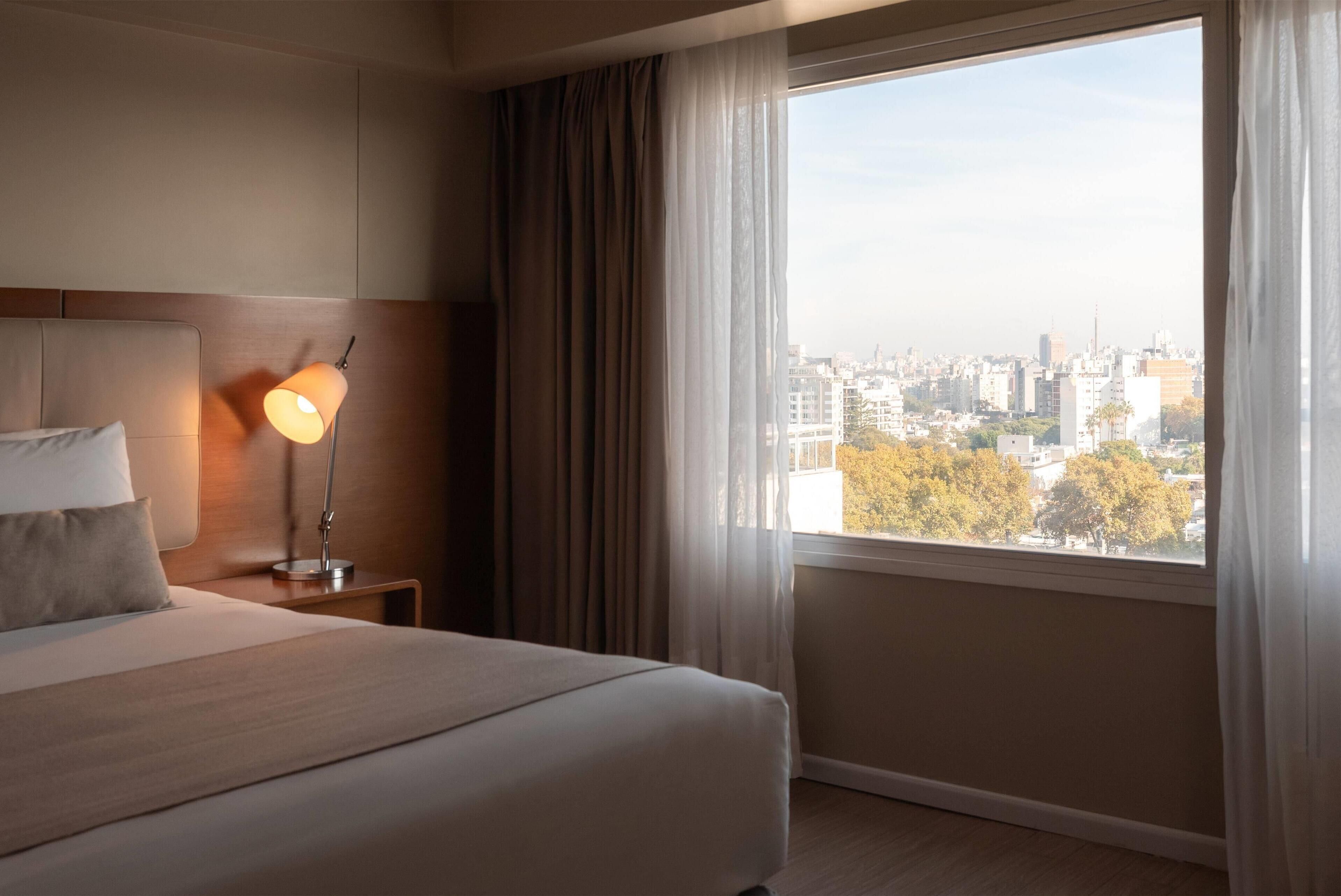 Habitación de hotel con una cama grande, una lámpara de noche y vistas a un paisaje urbano a través de un ventanal amplio.