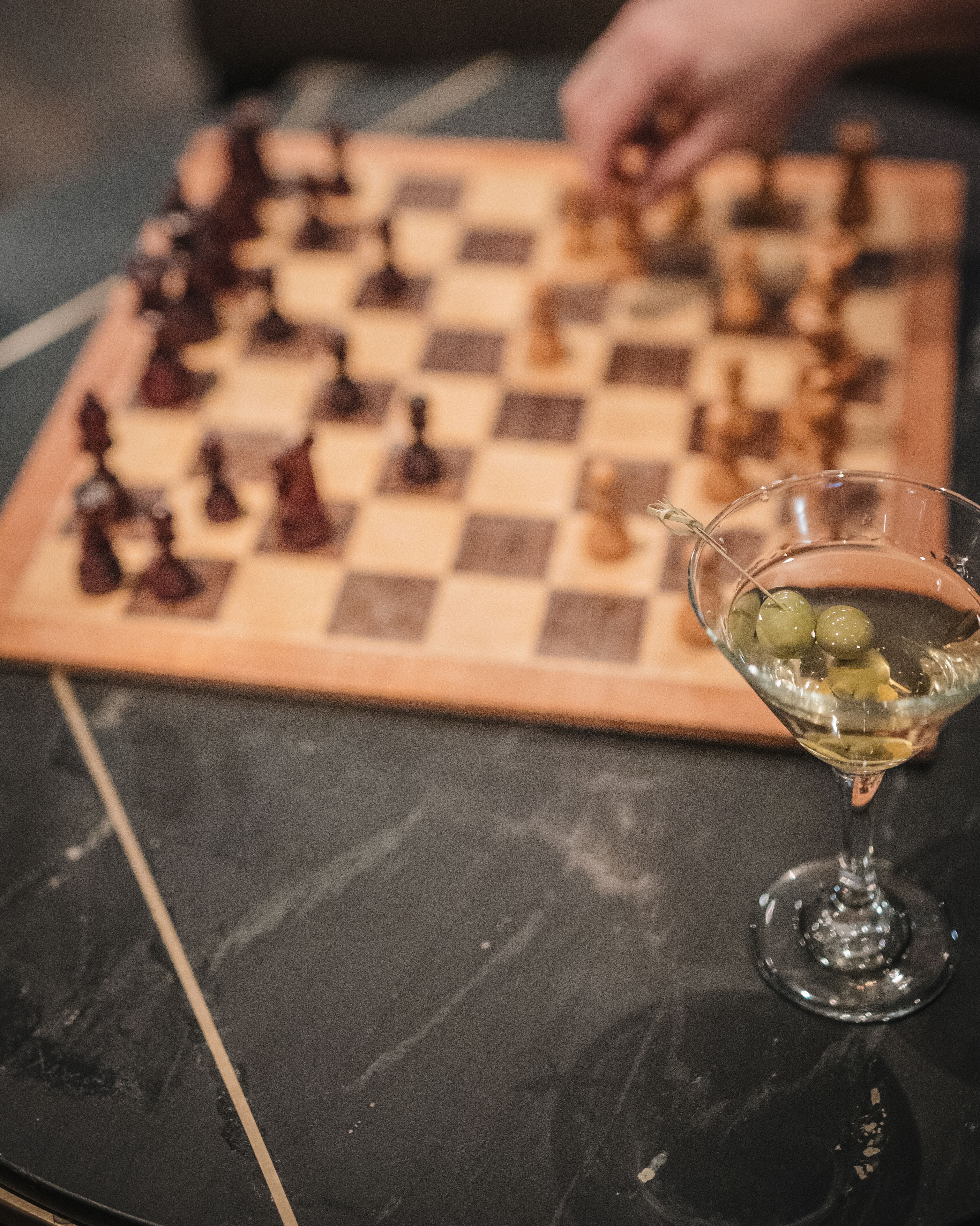 A chessboard with pieces and a martini glass with olives on a table.