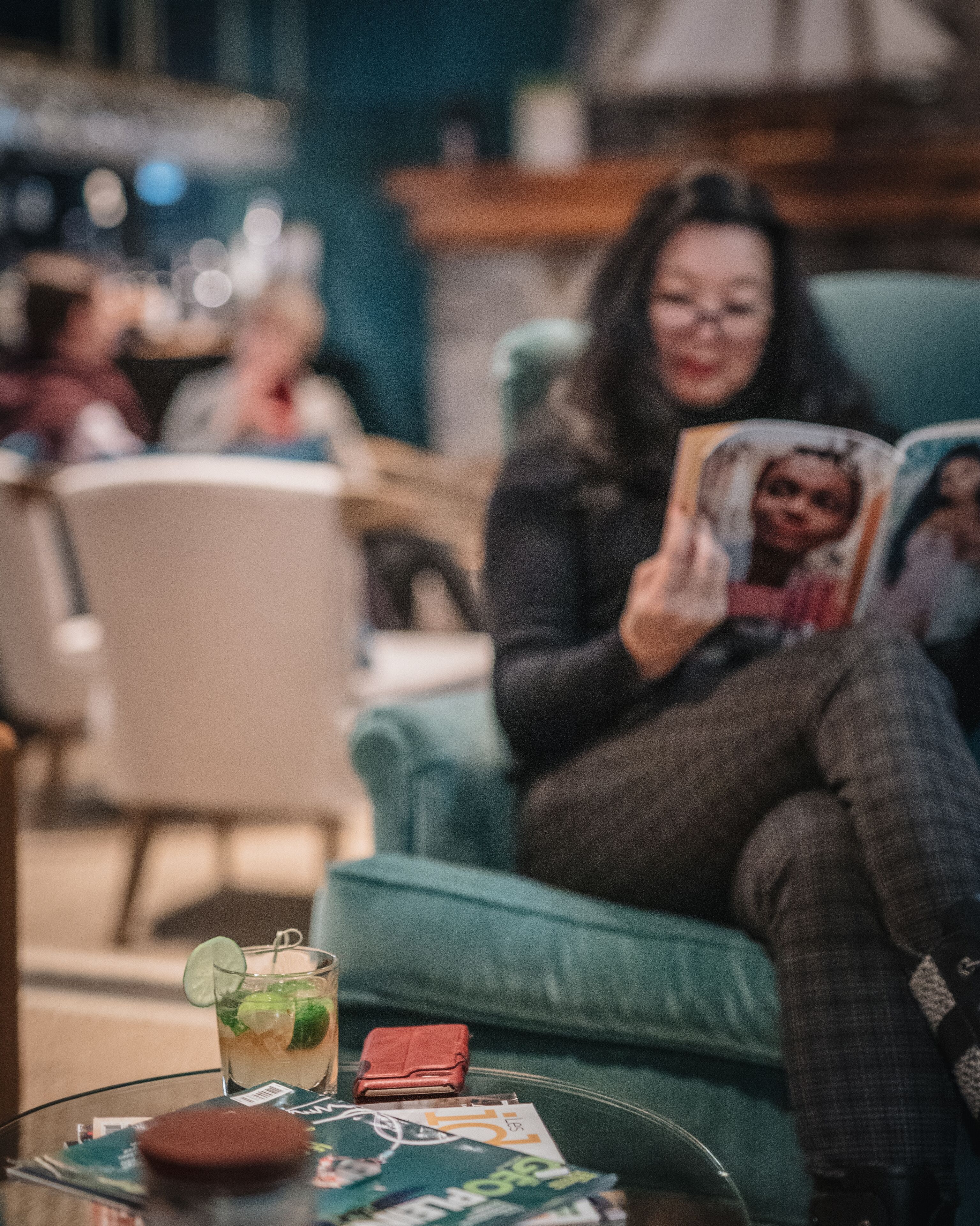 A person sitting on a teal armchair, reading a magazine. A glass table with a drink and a wallet is in front of them.