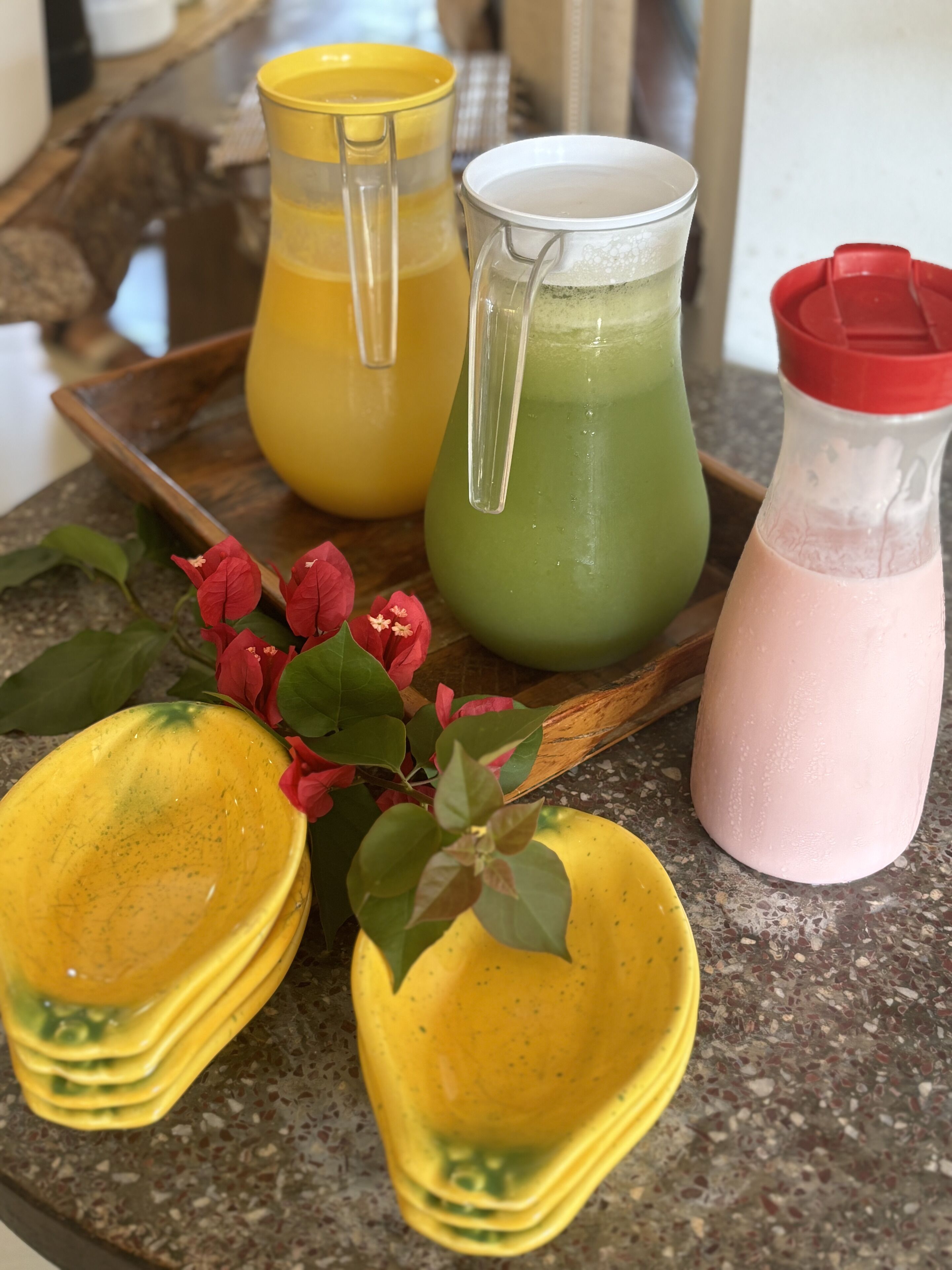 Una bandeja con tres vasos de bebidas de colores distintos, una pila de platos amarillos y una flor roja.